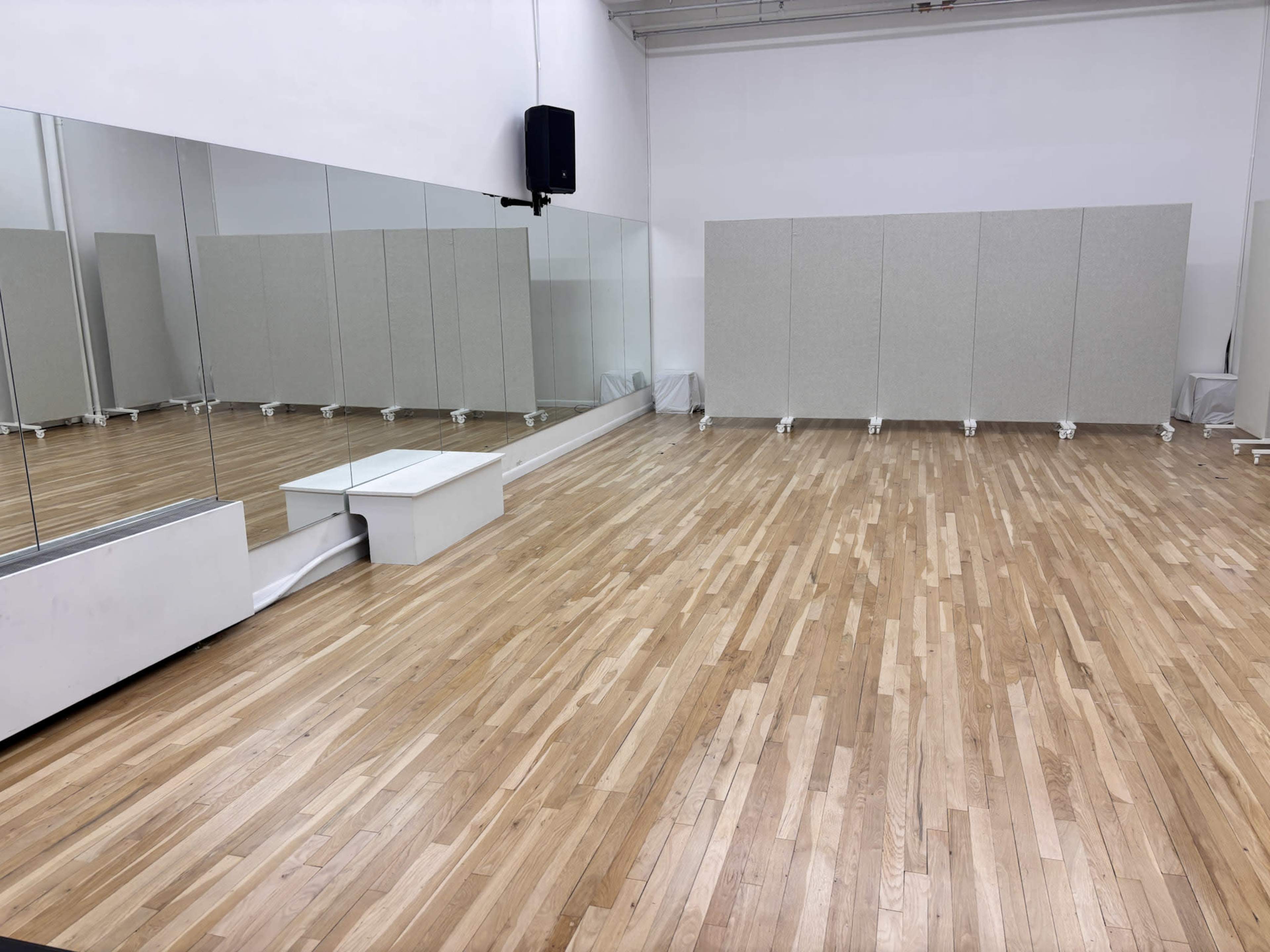 The image shows a spacious room with wooden flooring, large mirrors on one wall, and several movable partition panels set up to create separate areas.