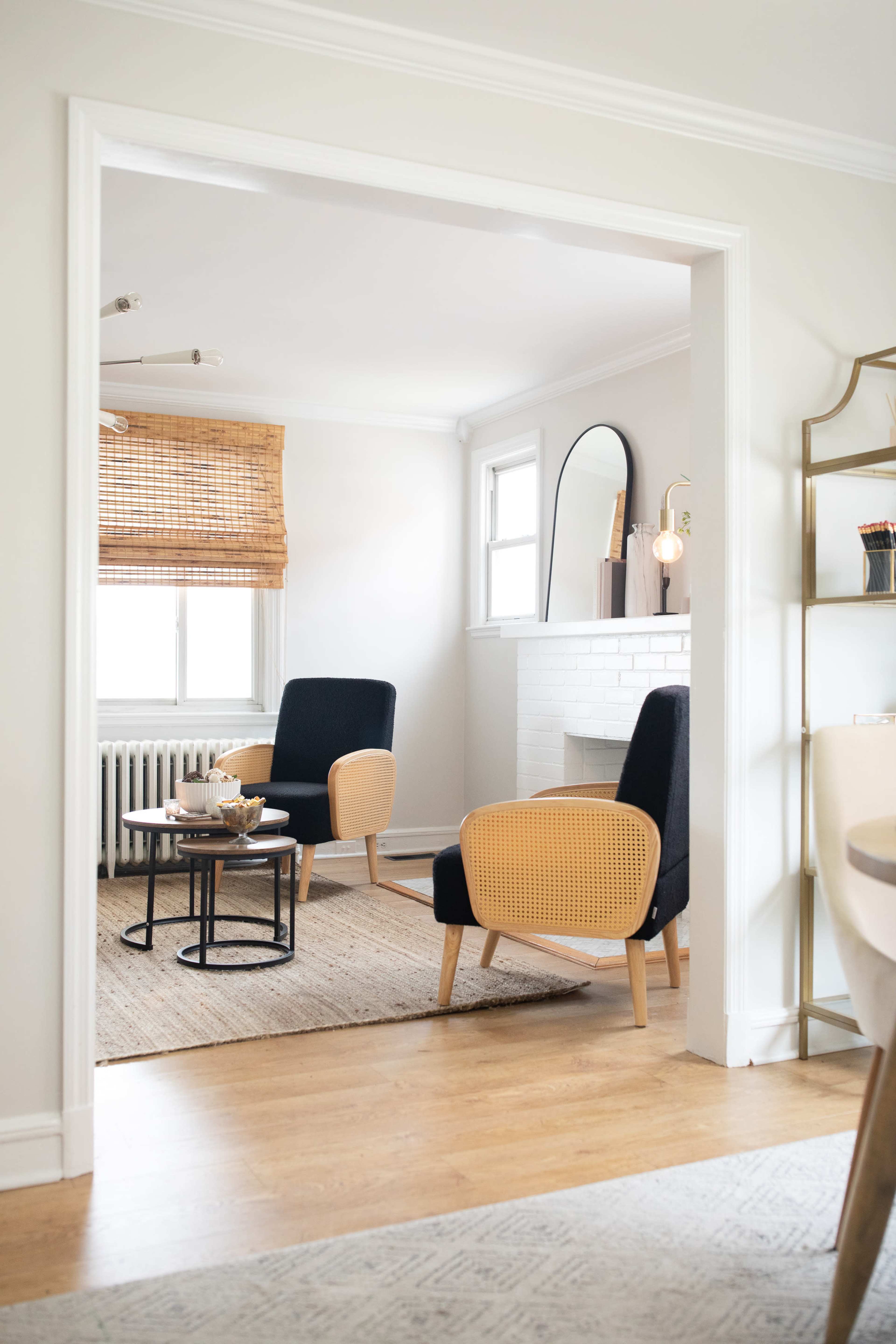 A bright room features two cane-back chairs and a black side table arranged around a central area rug, with a fireplace and window covered by bamboo shades in the background.