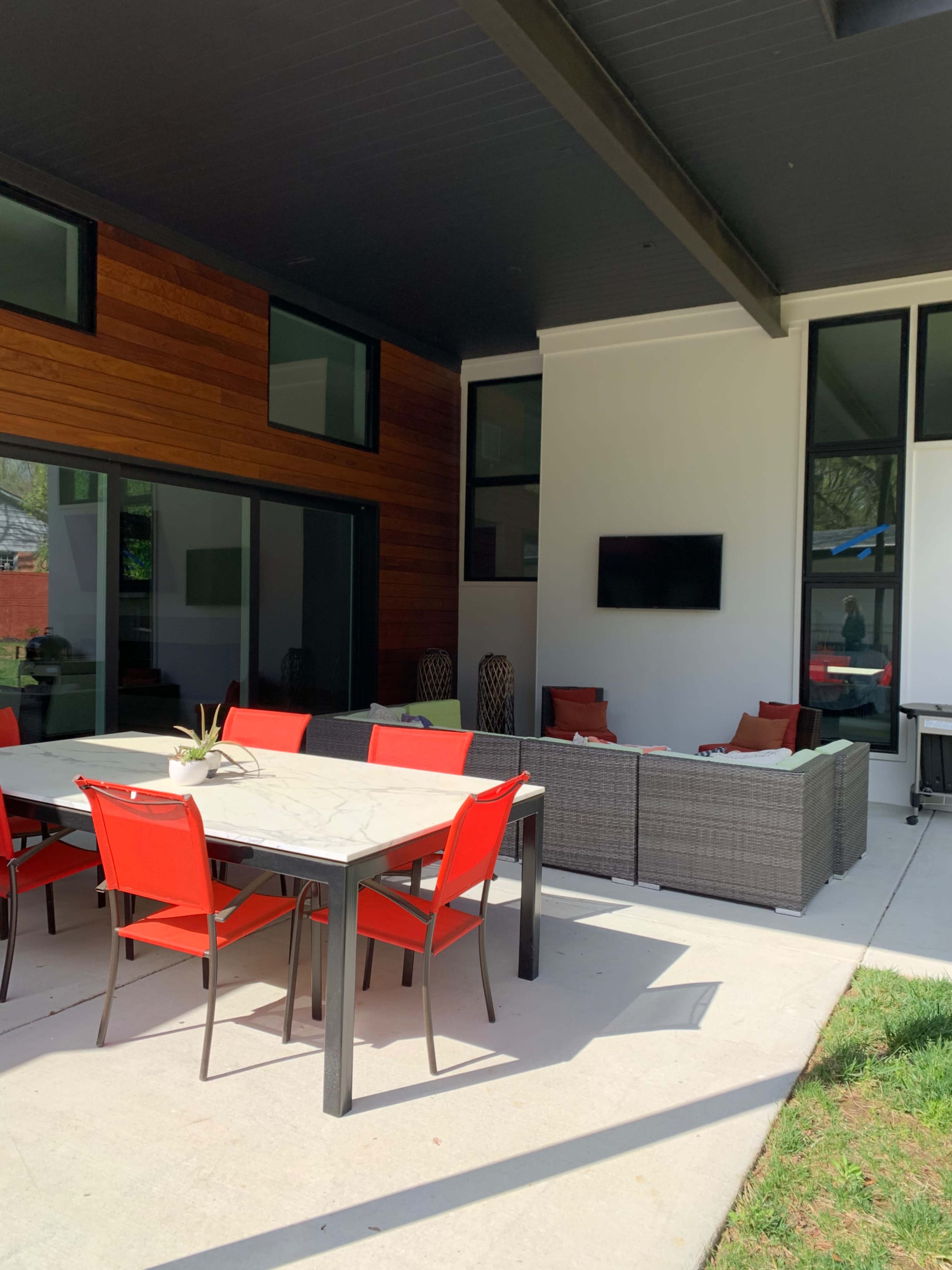 A modern outdoor patio features a dining table with red chairs and a seating area with a woven couch under a roofed structure.