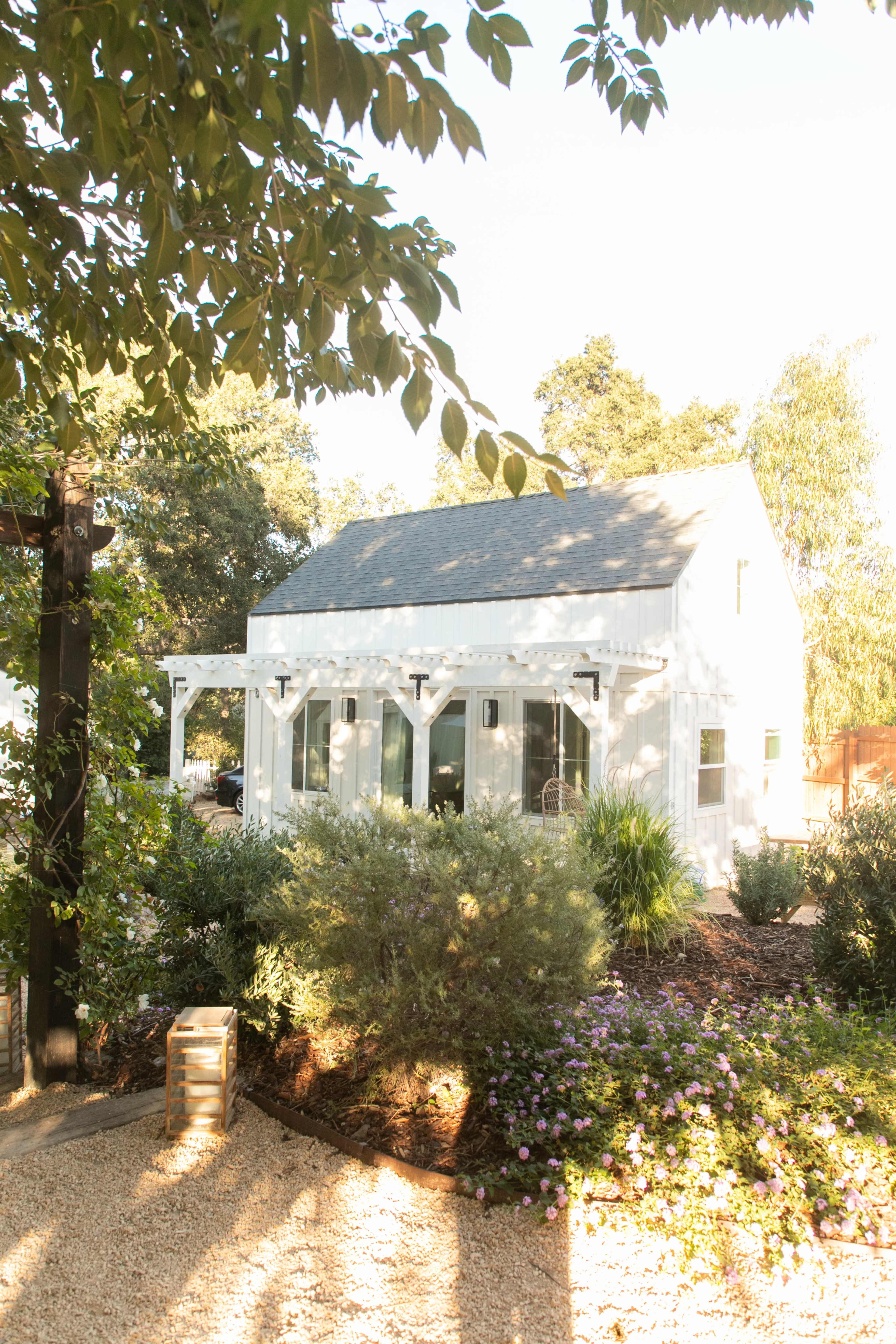 A small white house with a peaked roof is surrounded by greenery and a gravel pathway.