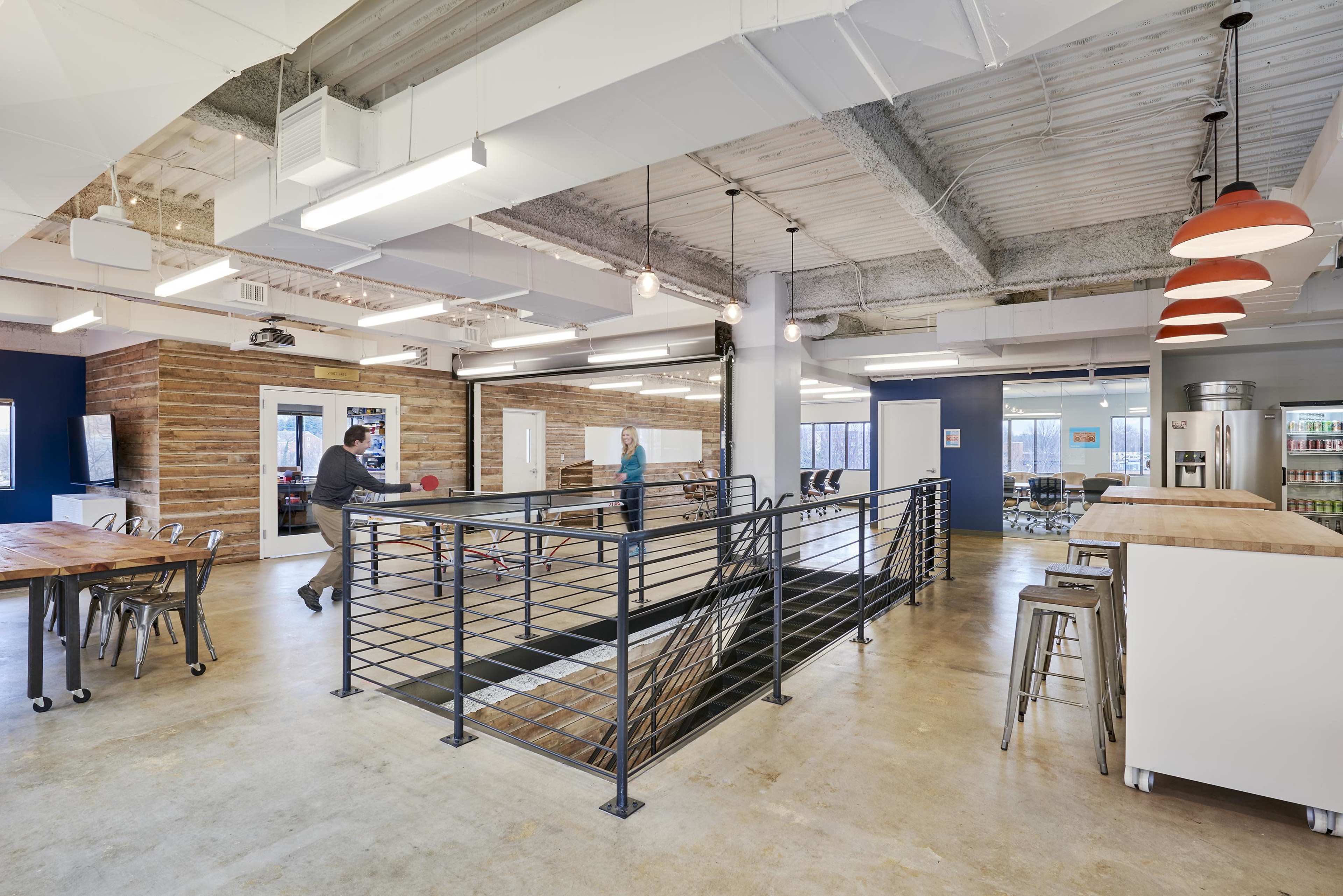 A modern open office space features a ping pong table, wooden tables, and a staircase leading to a lower level, with varied seating arrangements and industrial lighting.
