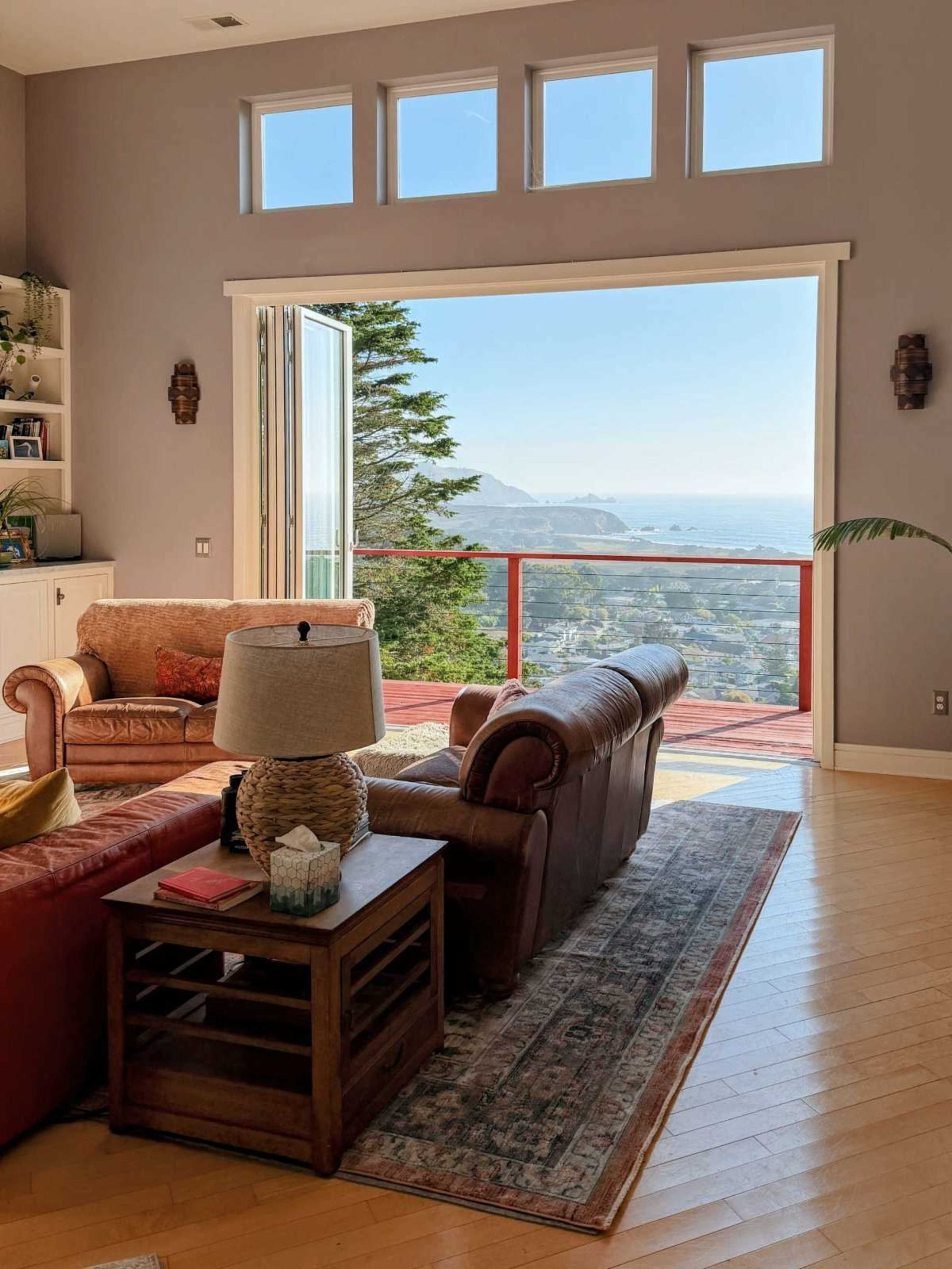 The living room features large open doors that lead to a scenic view of the coastline, with two sofas and a coffee table positioned on a wooden floor.