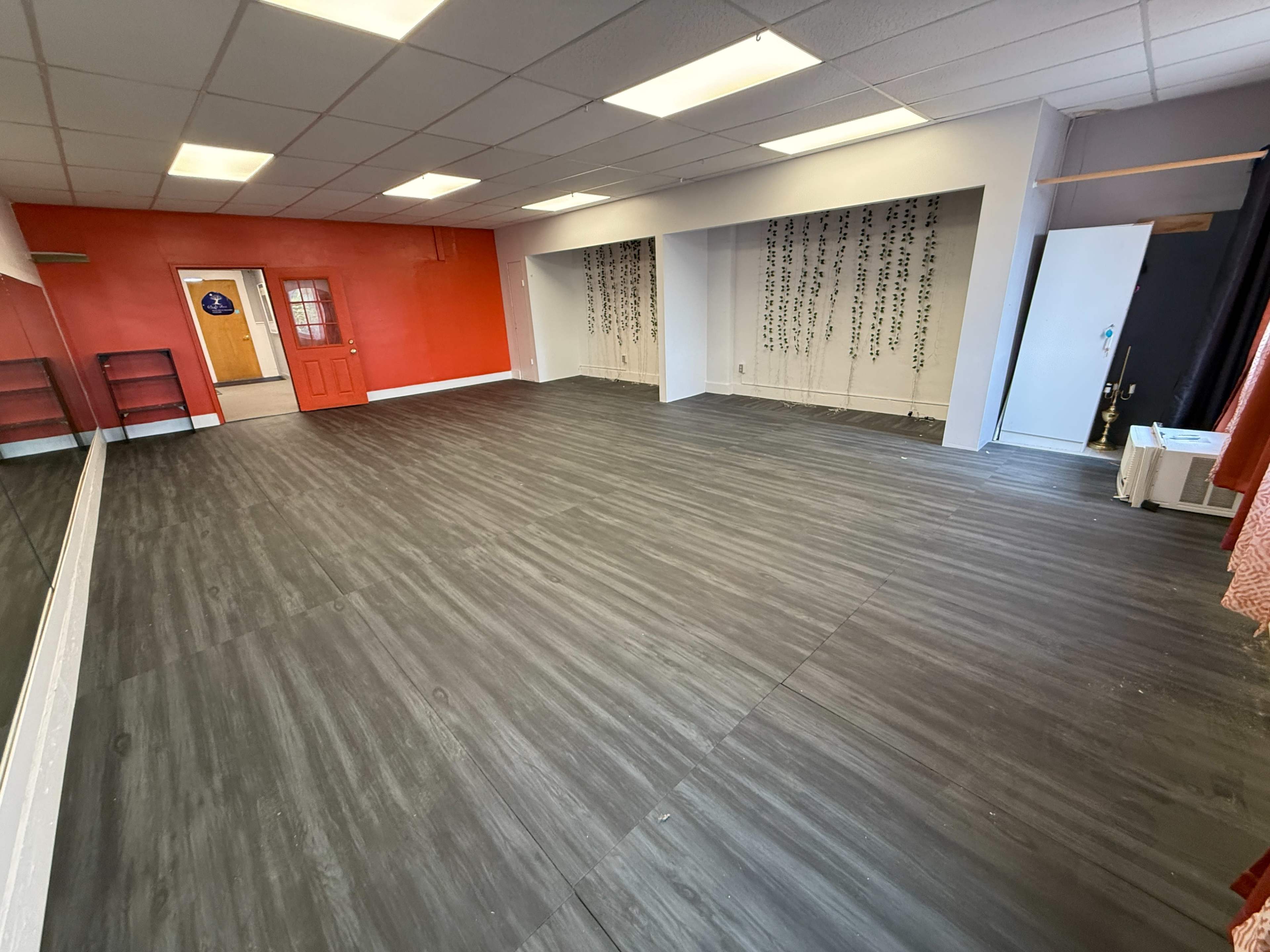 The image shows a spacious indoor room with gray flooring, red accent walls, and minimal furnishings including a few shelves and a doorway leading to another area.