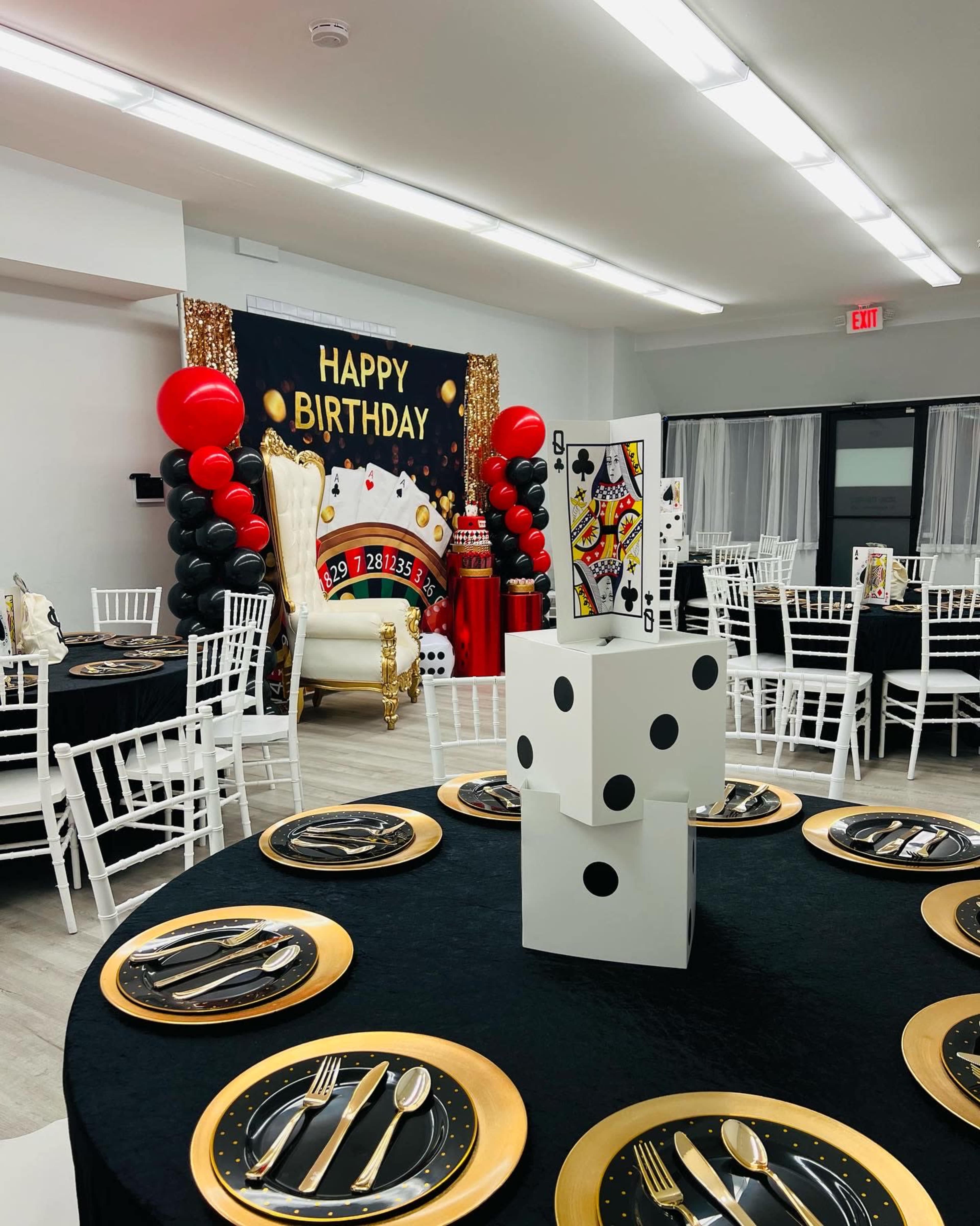 The image shows a decorated birthday party venue with a black and gold theme, featuring a large birthday banner, balloons, and a cake table adorned with dice and playing card graphics.
