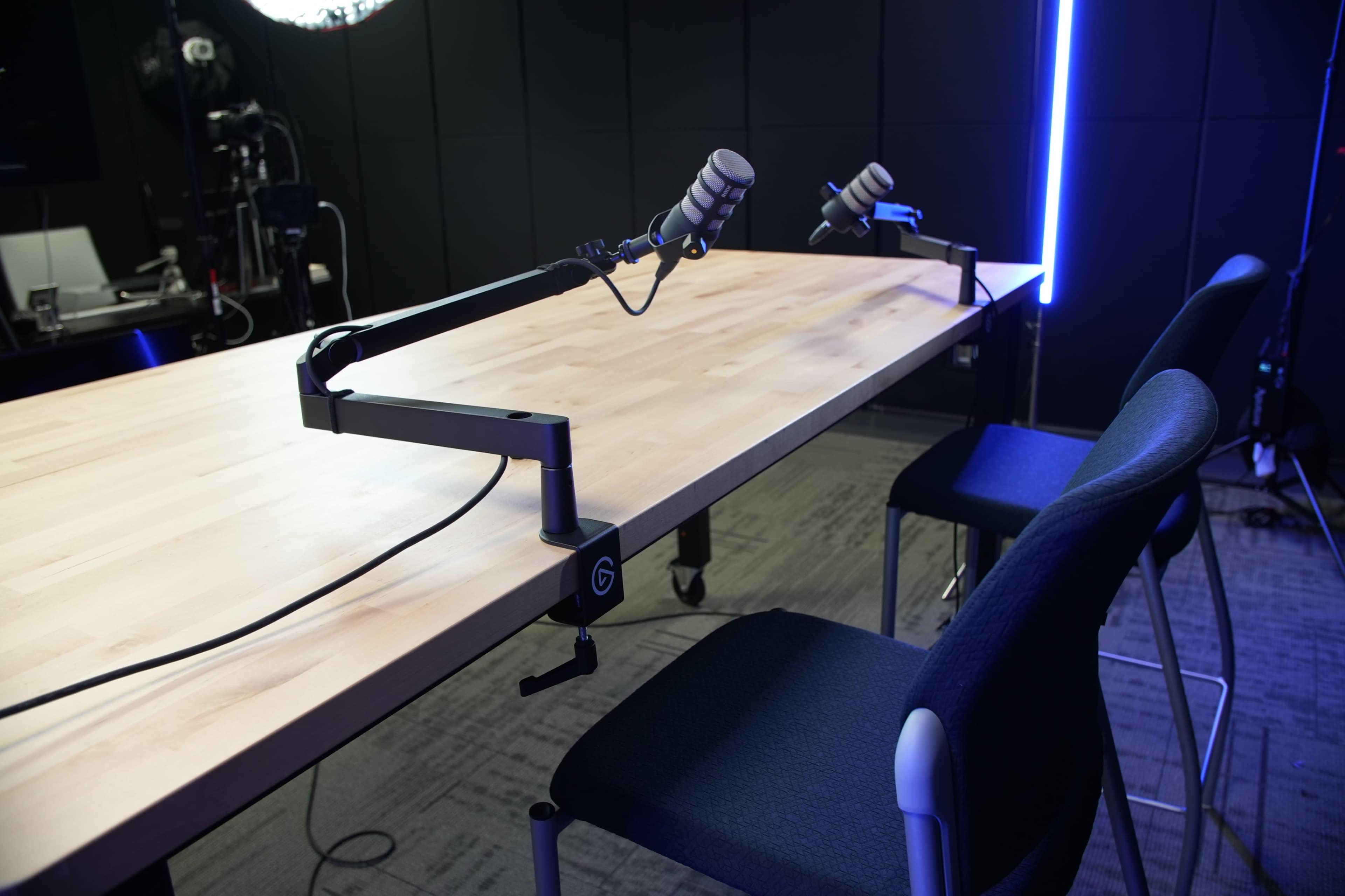 A wooden table with two microphone mounts and two empty chairs is set up in a recording studio.