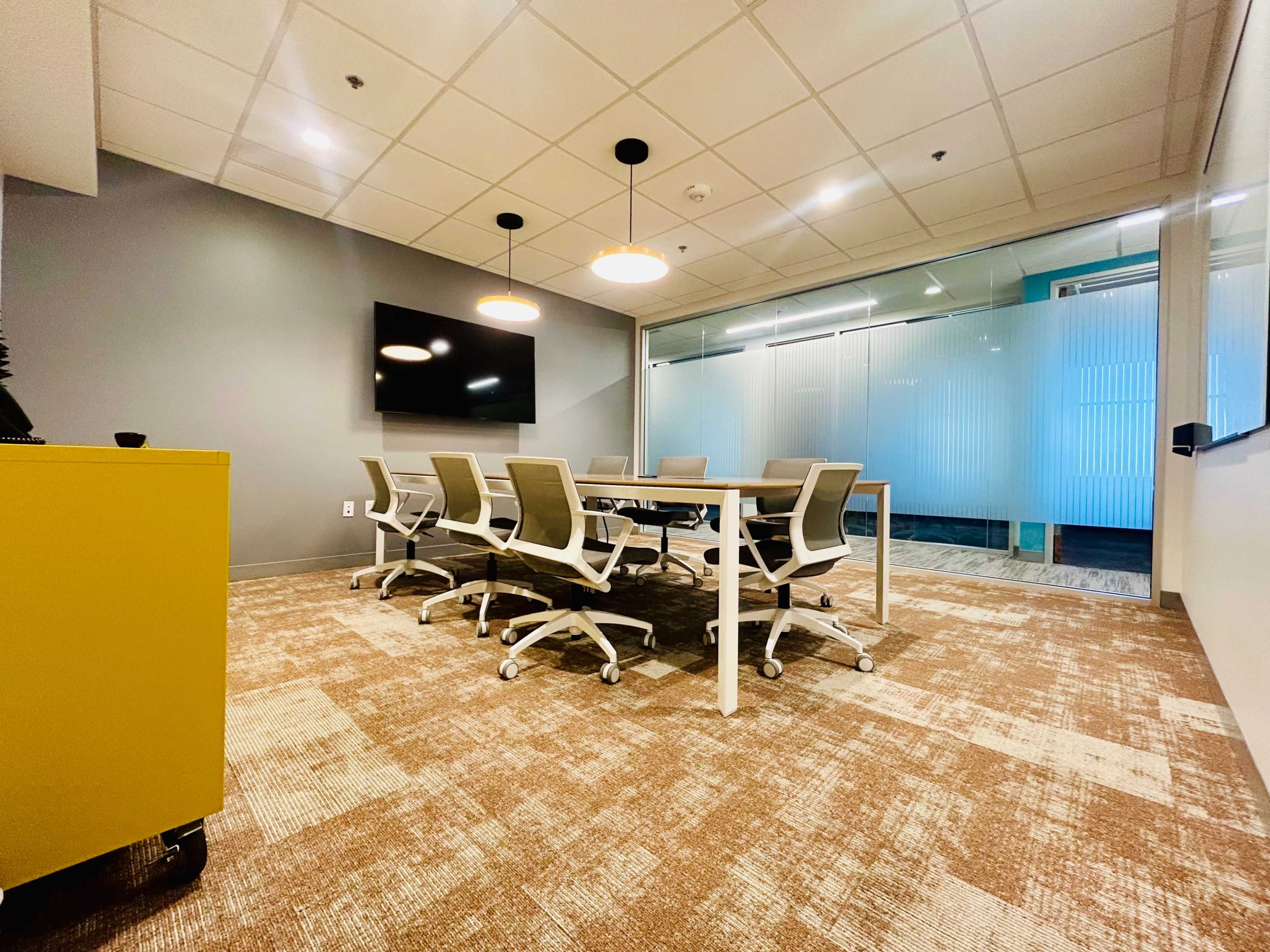 A modern conference room features a long table surrounded by ergonomic chairs, a large wall-mounted screen, and glass partitions allowing natural light to enter.