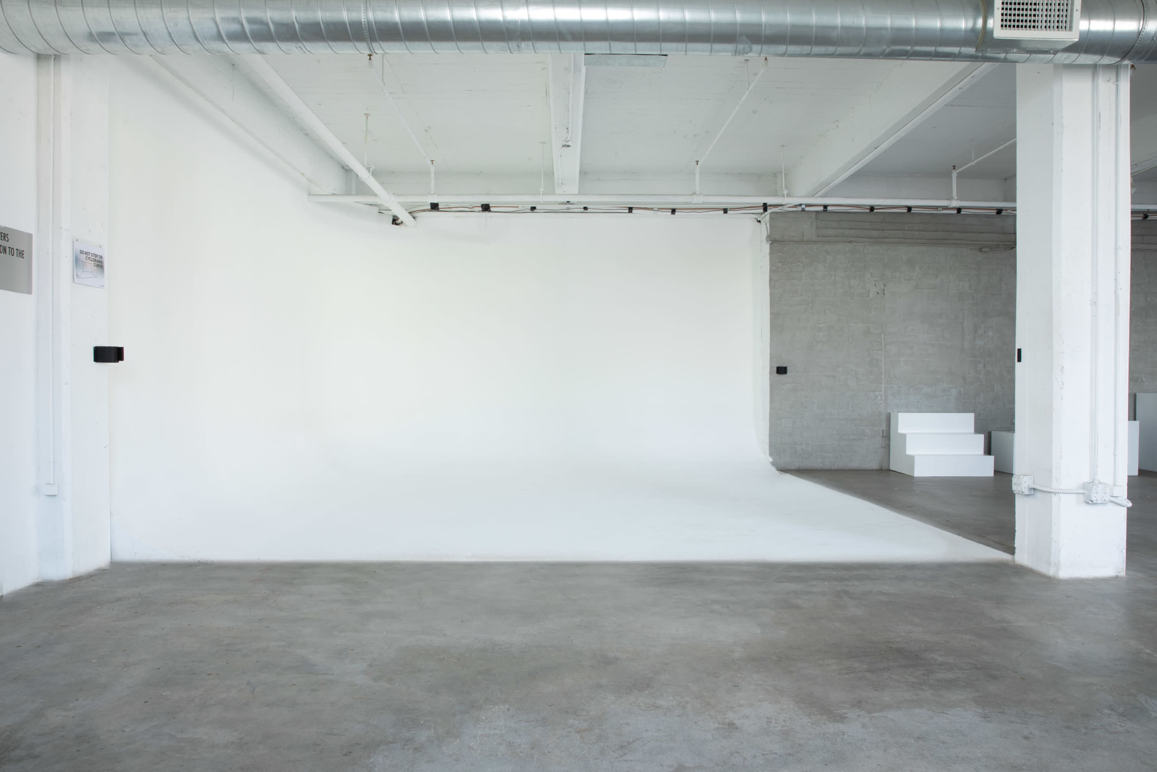 The image shows a spacious, empty room with a white backdrop and concrete flooring.