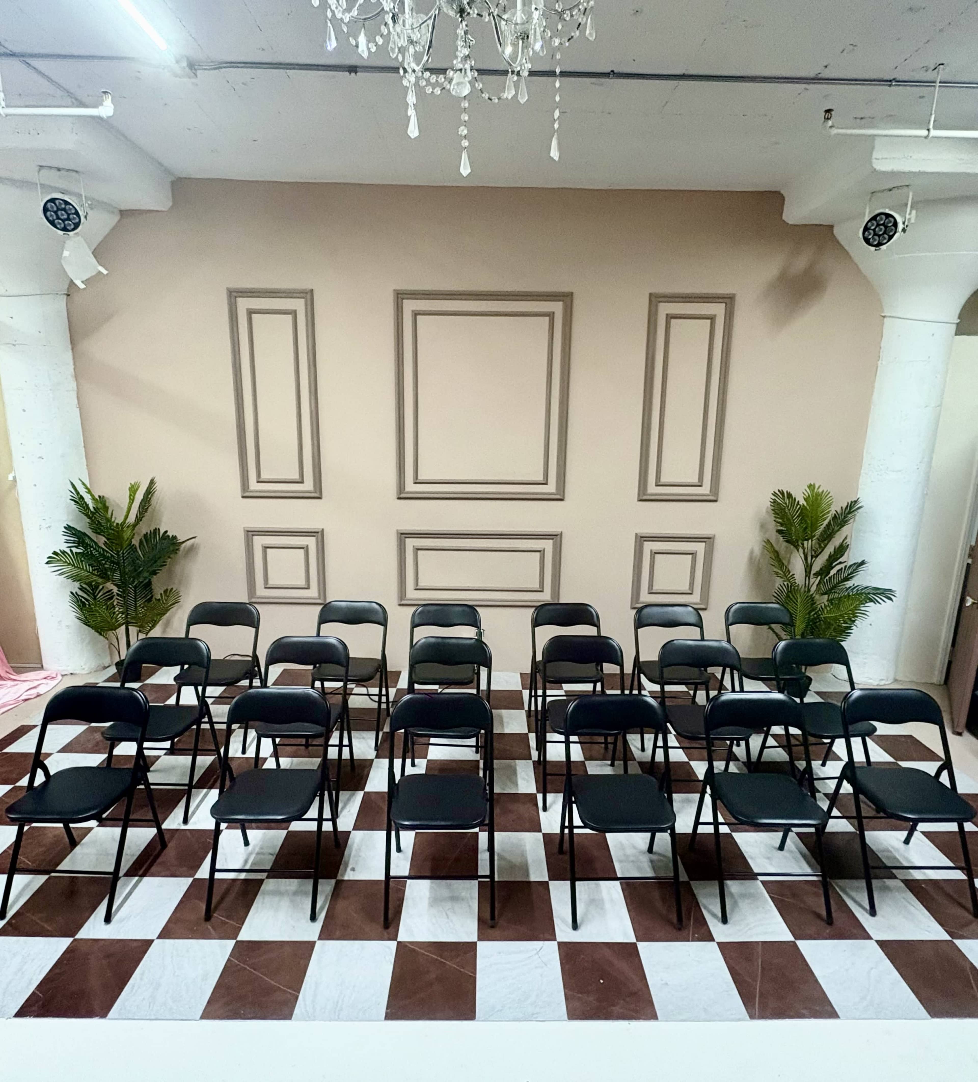 The image shows a setup of black folding chairs arranged in rows on a checkered floor, with a neutral-colored wall featuring decorative paneling and two potted plants in the corners.