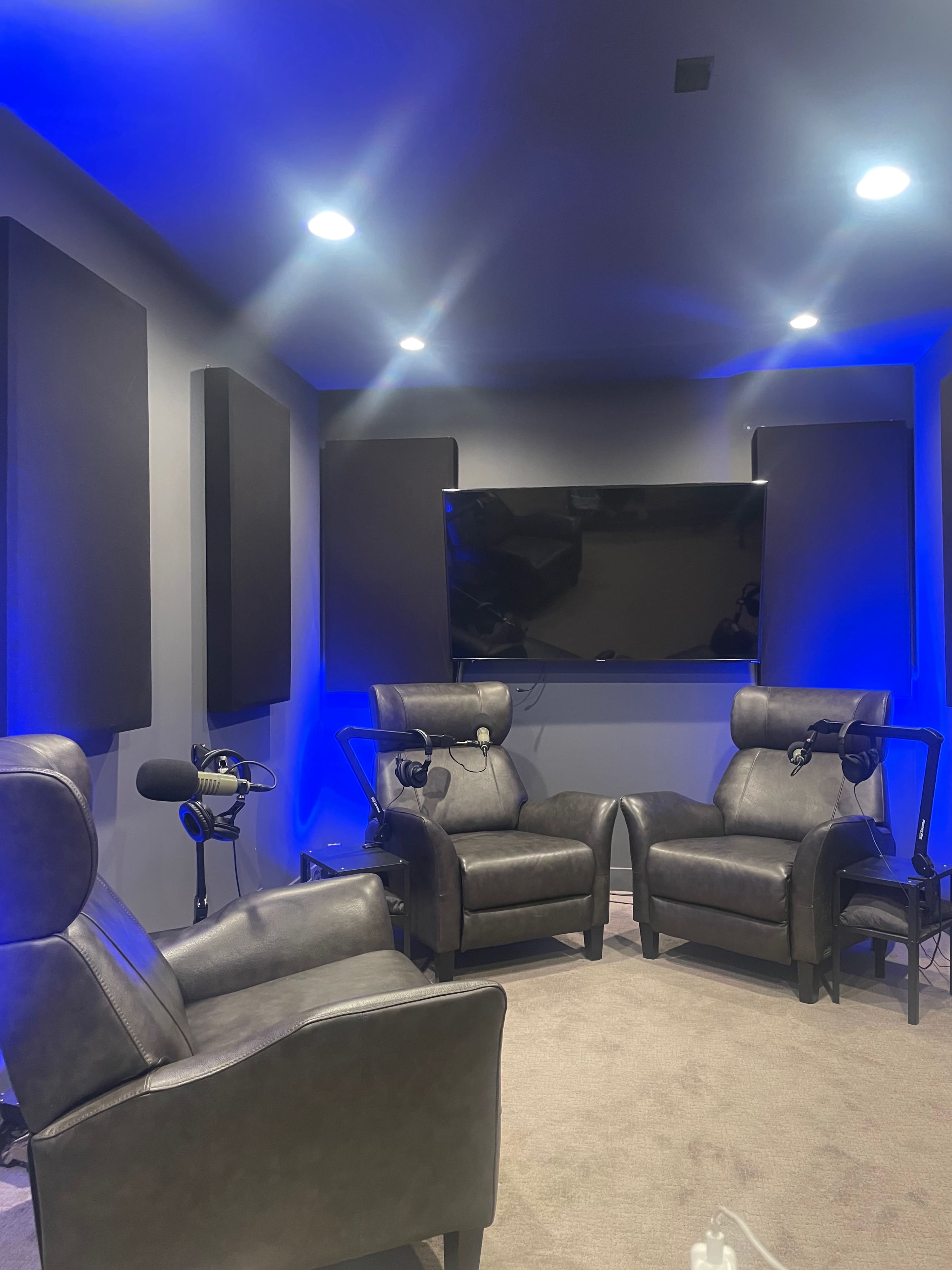 A small recording studio featuring two leather chairs, a television mounted on the wall, and soundproof panels on the sides, illuminated by blue lighting.