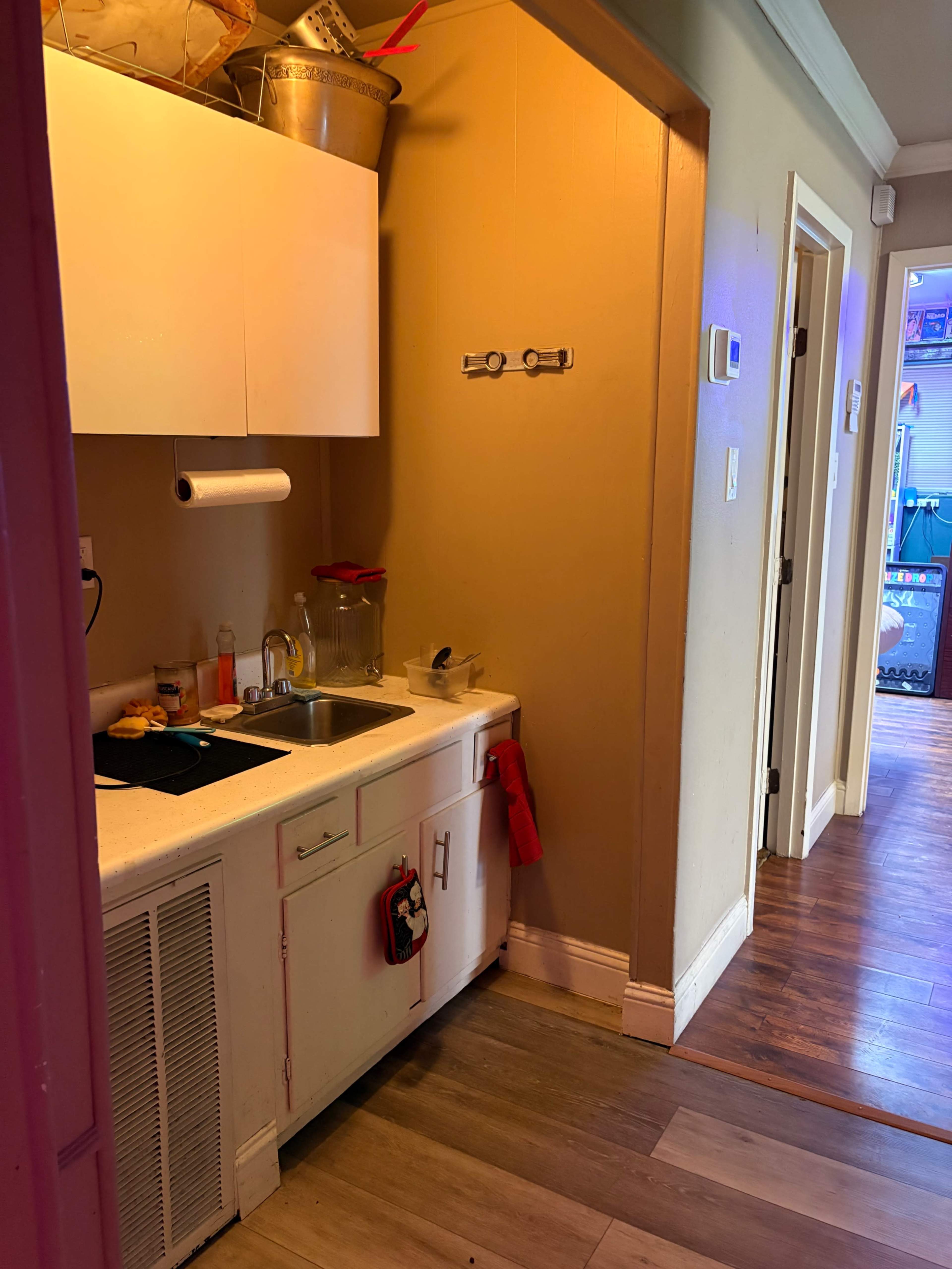 A small kitchenette with a sink and countertop is situated at the end of a hallway, featuring light-colored walls and hardwood flooring.