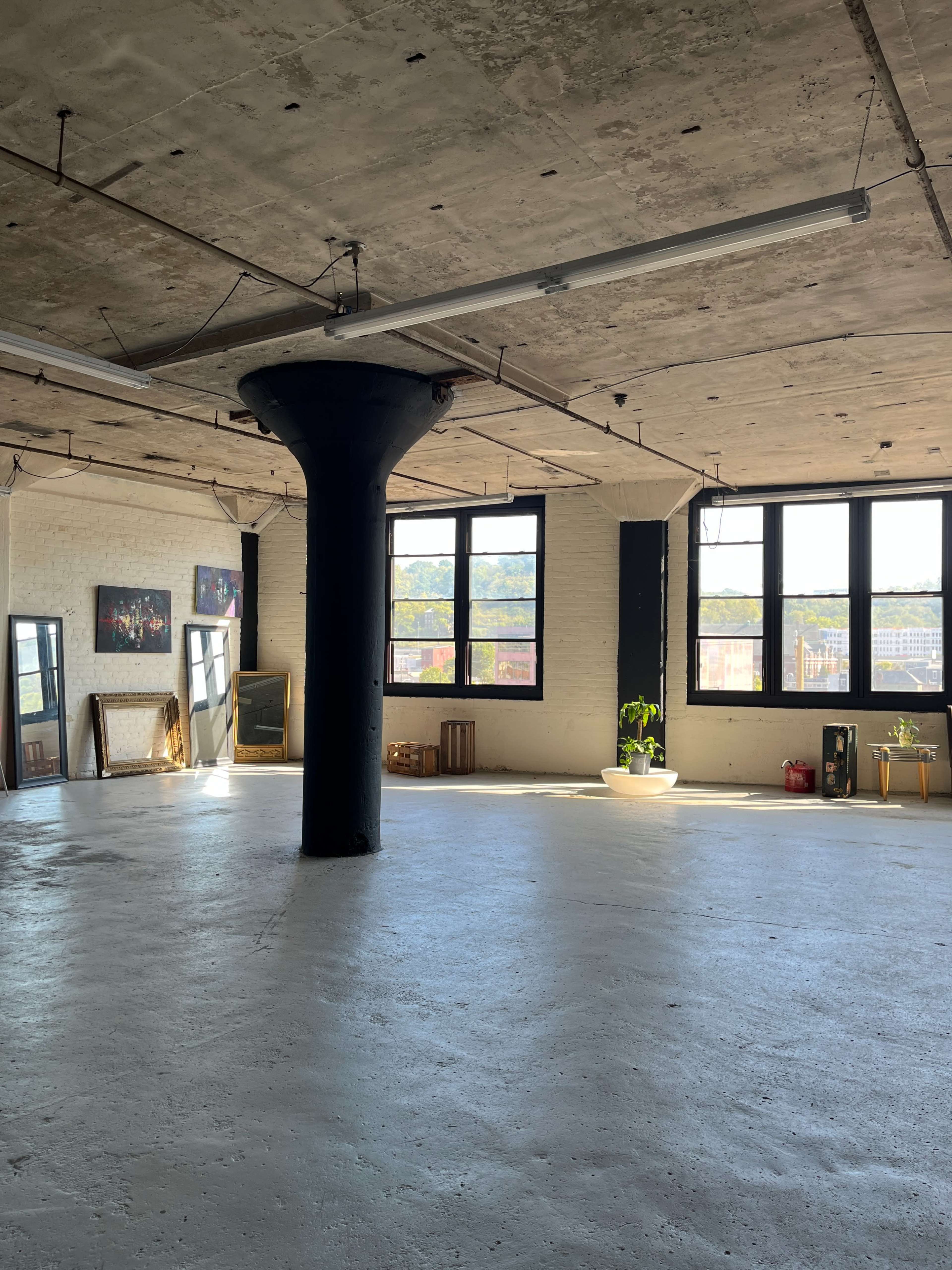 An empty industrial-style room with large windows, a concrete floor, and a central black column.