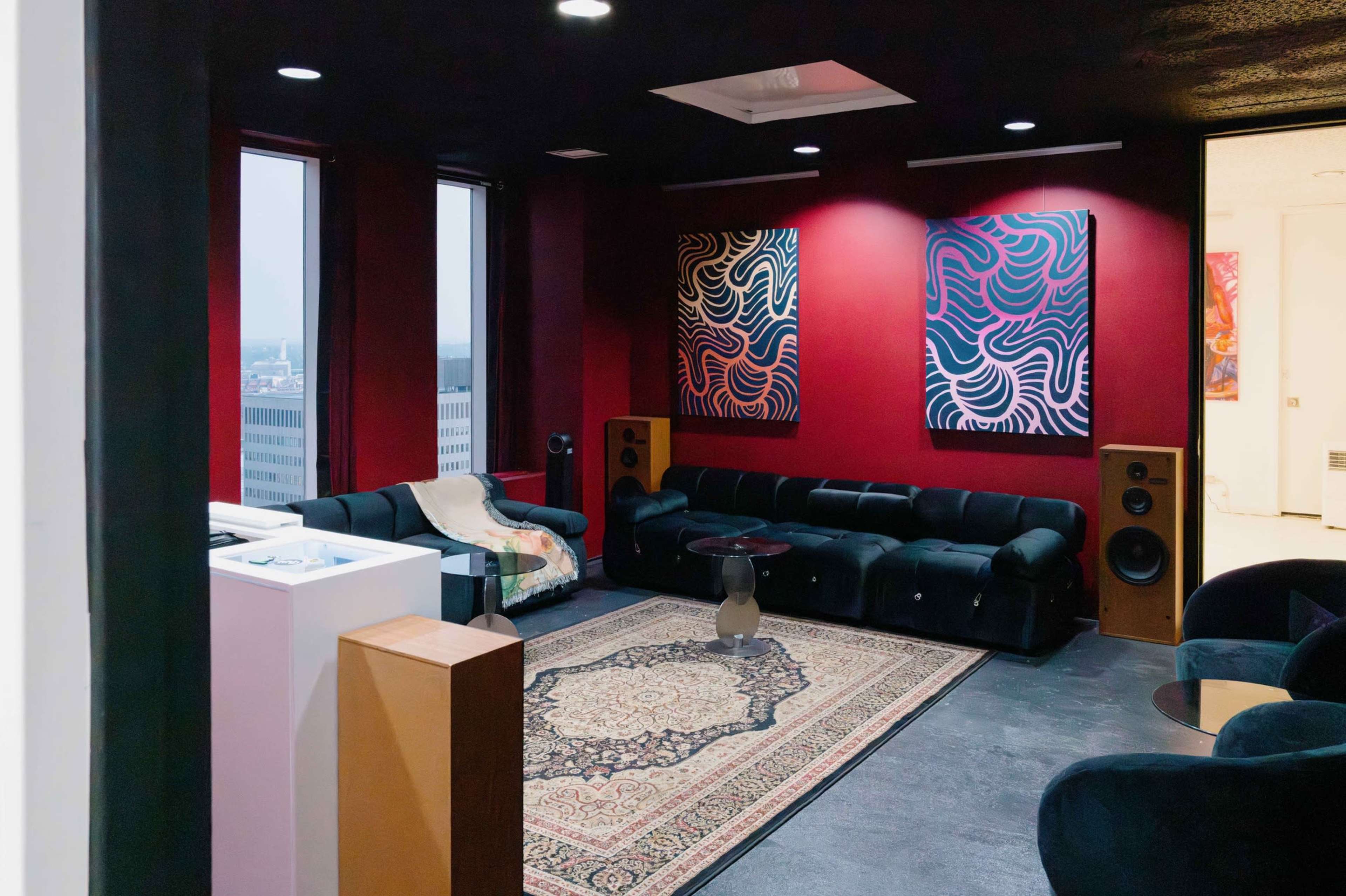 A modern lounge area features black sofas, a round glass table, colorful abstract artwork on the walls, and large windows overlooking a cityscape.