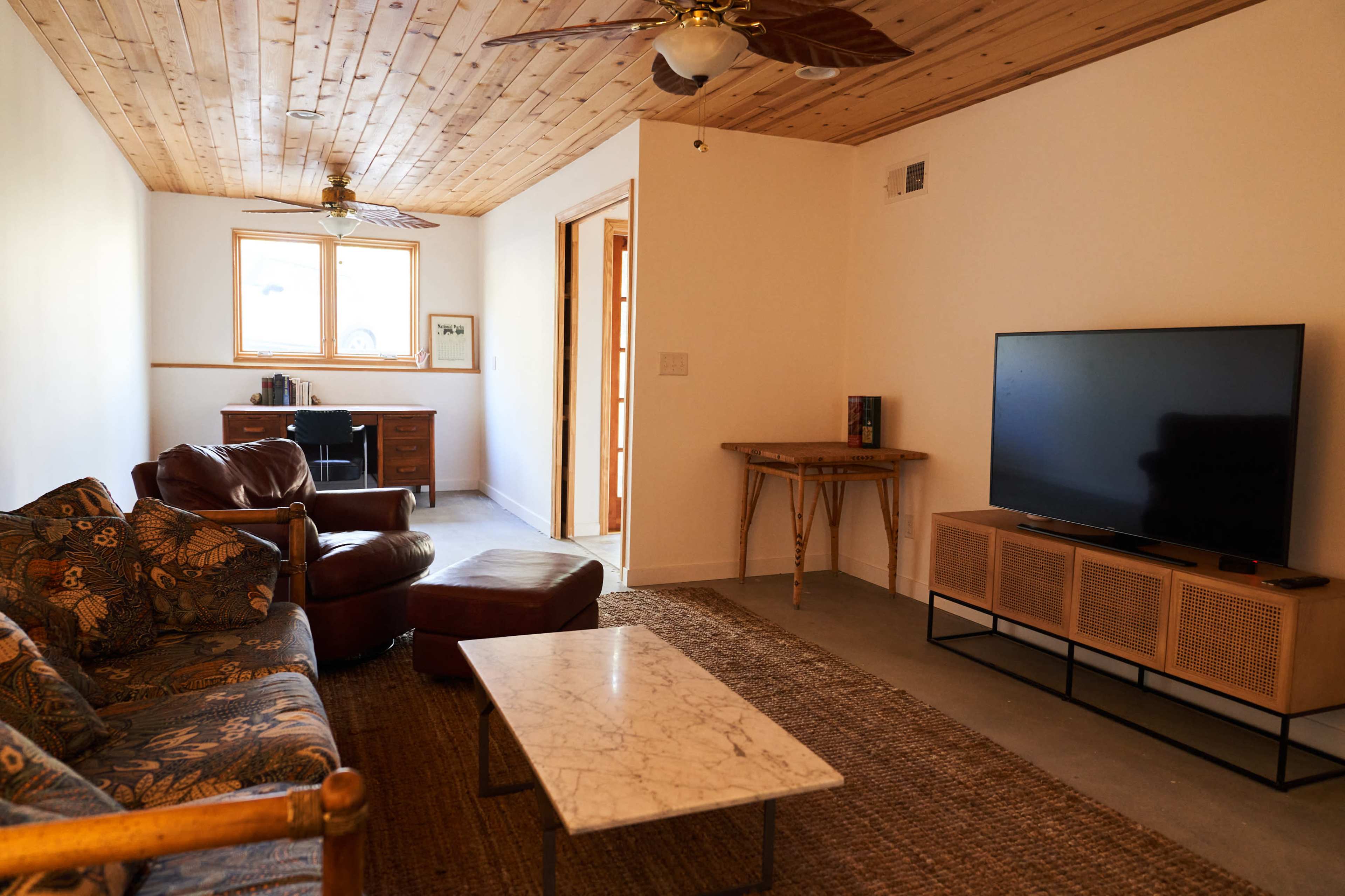 A living room features a sofa, armchair, coffee table, and a television on a stand, with a desk visible in the background and natural light coming from a window.