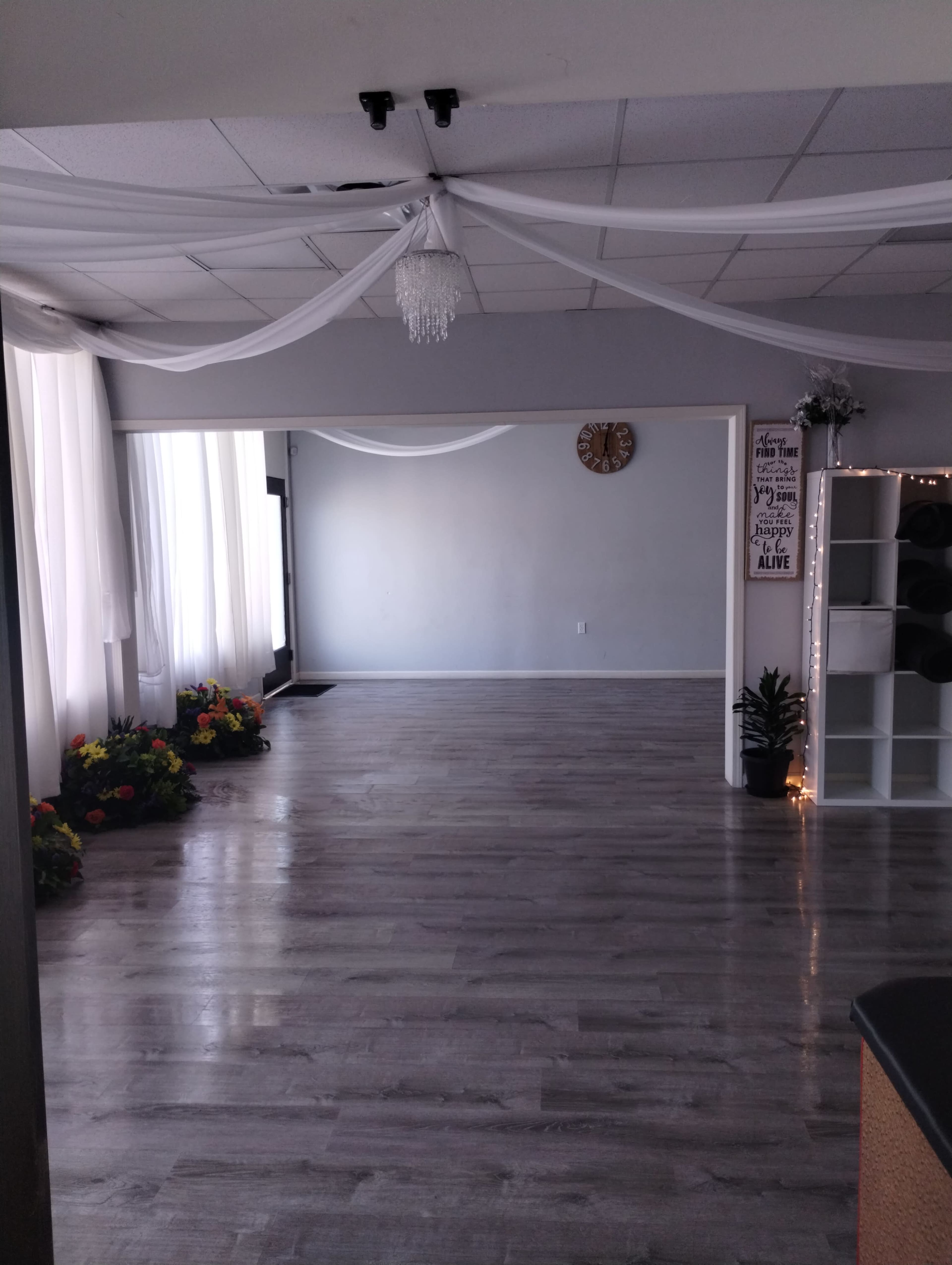 A spacious studio with wooden flooring, large windows draped with white fabric, and flower arrangements in the corners.