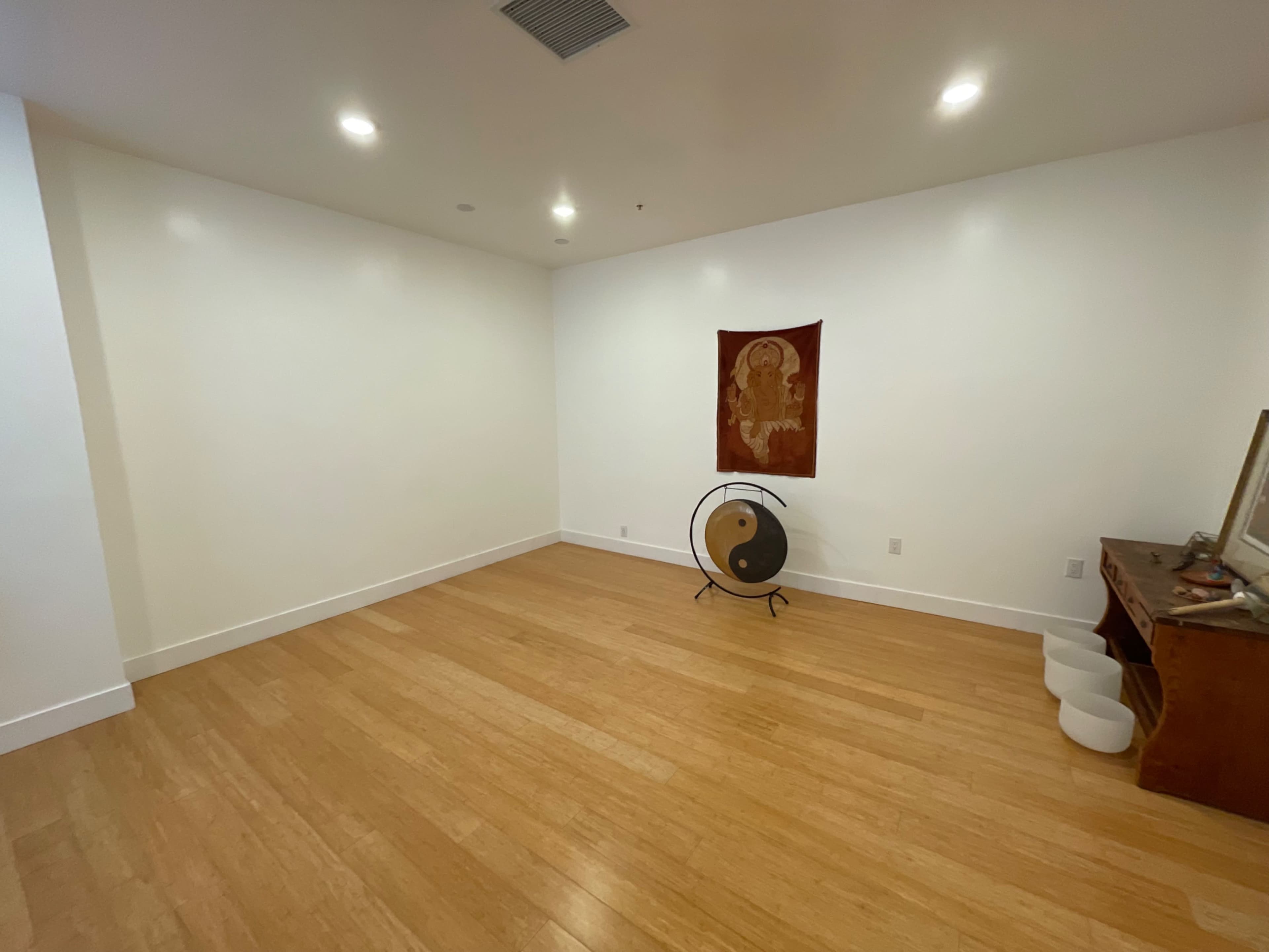 A sparsely furnished room with bamboo flooring, featuring a wall tapestry, a gong on a stand, and a wooden cabinet.