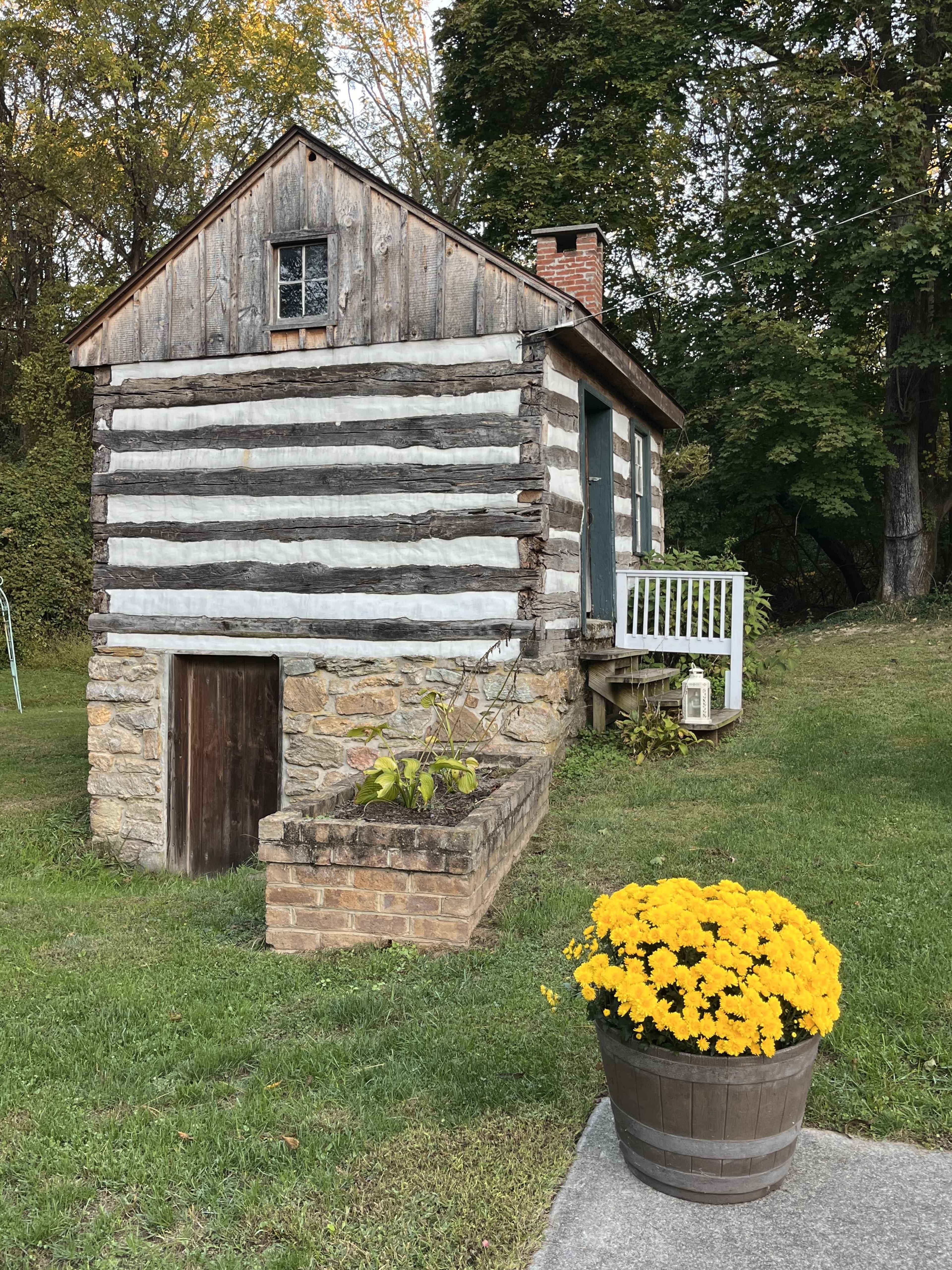 A small log cabin with a stone foundation sits in a grassy yard, accented by a pot of bright yellow flowers in the foreground.
