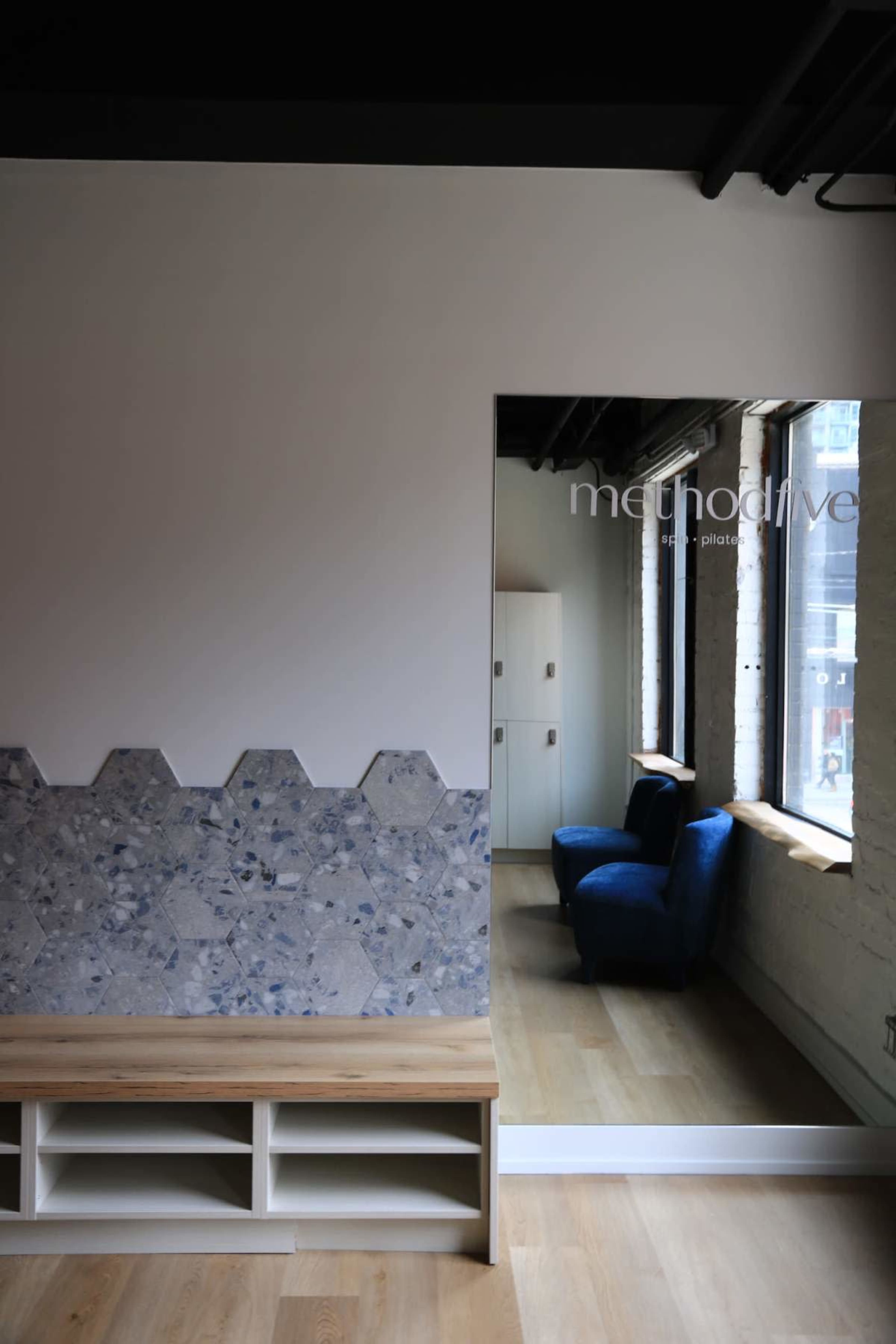 The image shows a modern interior space featuring a hexagonal stone accent wall, a wooden shelf, and a blue chair positioned near a large window.
