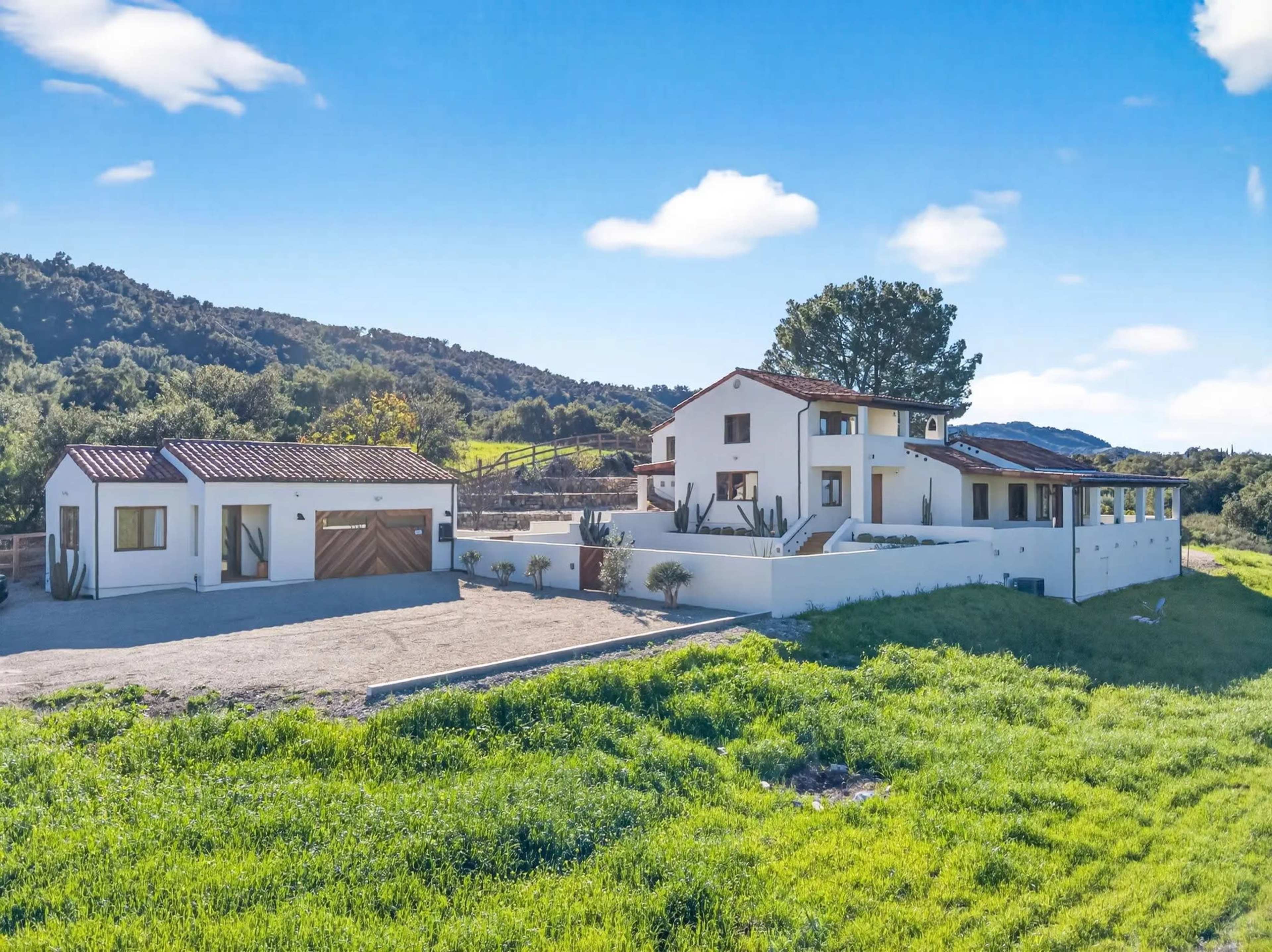 The image shows a modern farmhouse with a white exterior and red-tiled roof situated in a green, hilly landscape.
