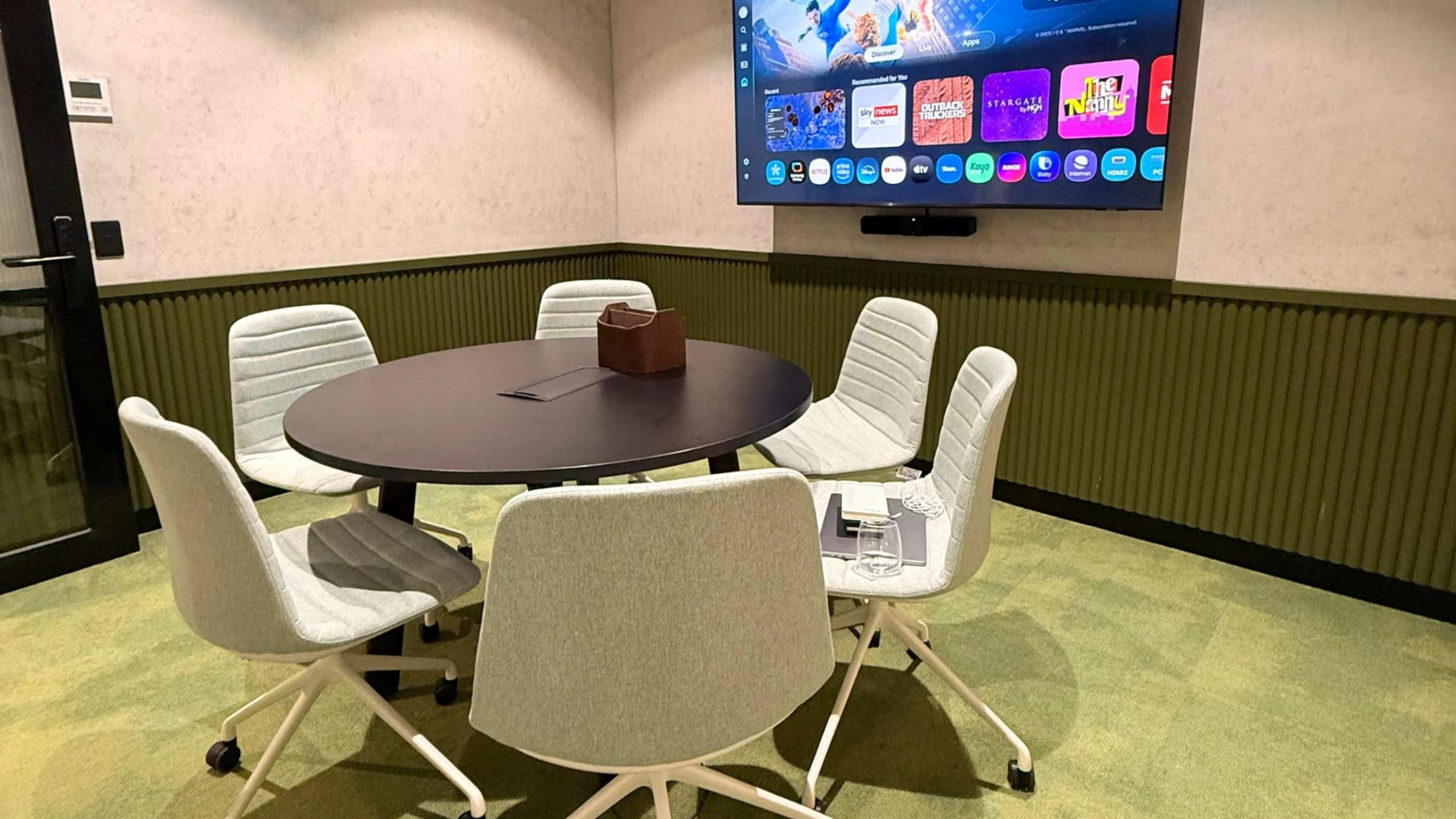 A modern conference room features a round table surrounded by five chairs and a large screen displaying various applications on the wall.