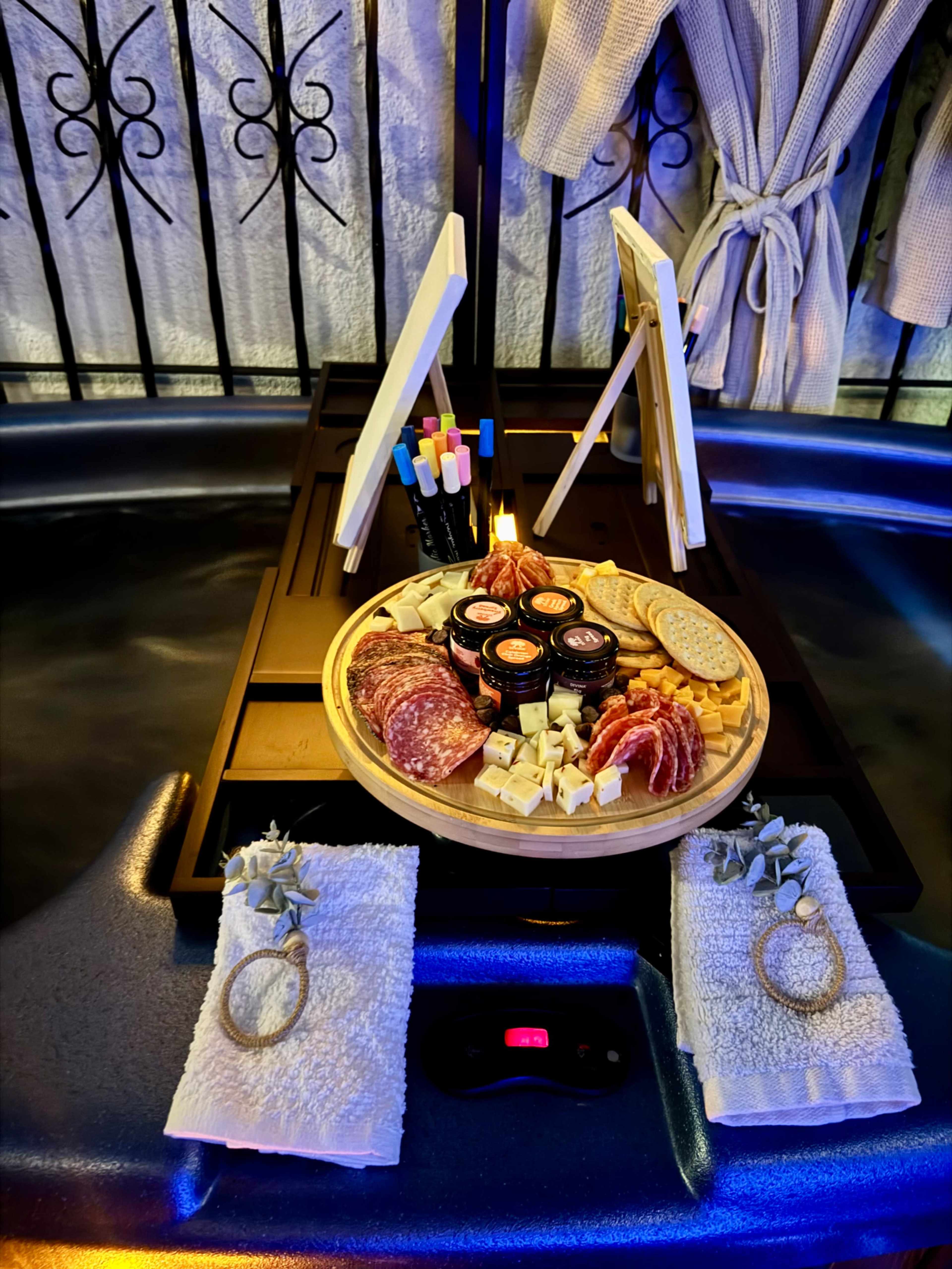A wooden platter with various meats and cheeses is set on a table beside a hot tub, accompanied by towels and small easels with markers.