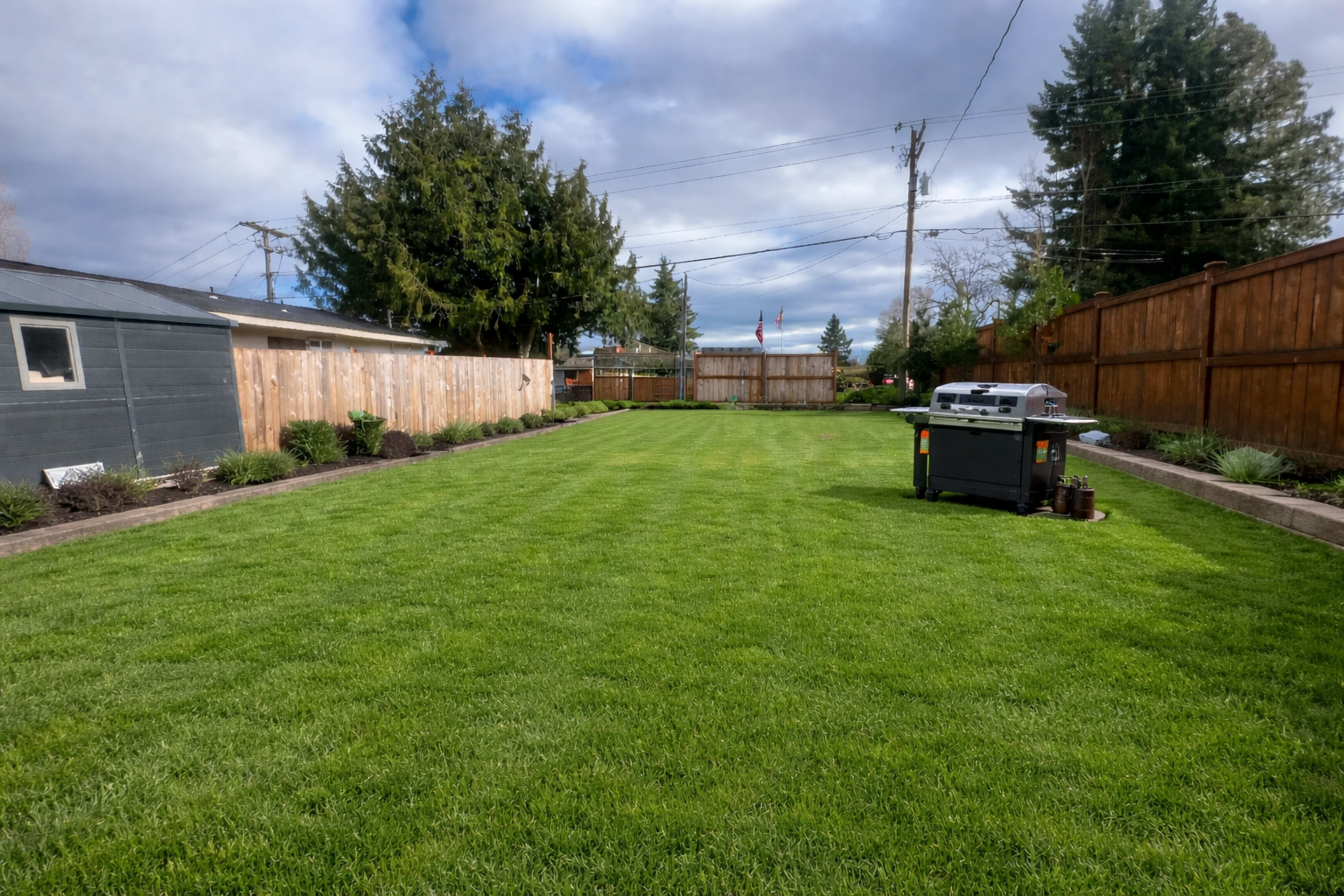 The image shows a neatly landscaped backyard with a green lawn, bordered by wooden fences and a small deck area on one side.