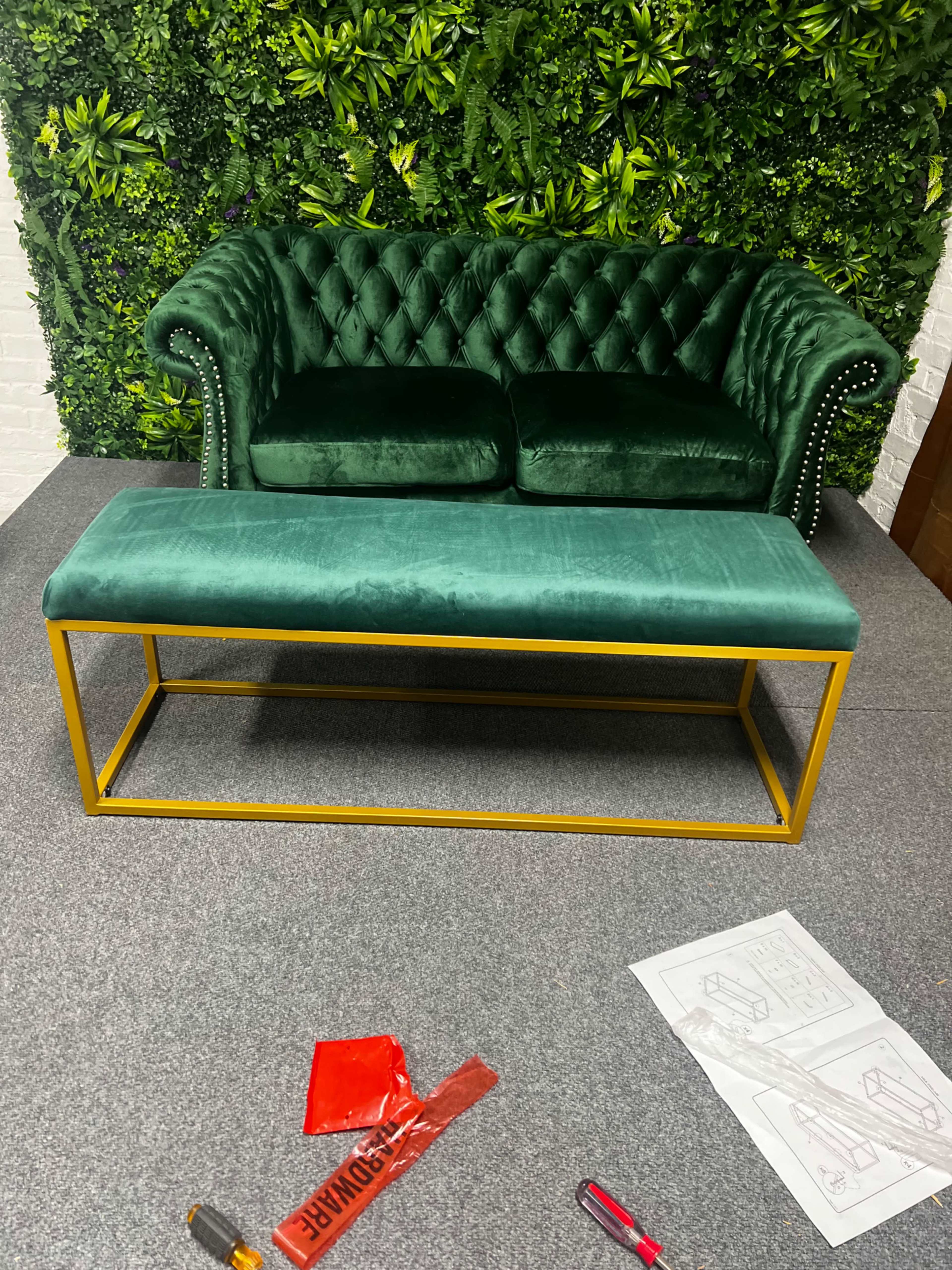 A green velvet loveseat with tufted upholstery is placed on a gray carpet in front of a gold metal coffee table and surrounded by a wall of greenery.