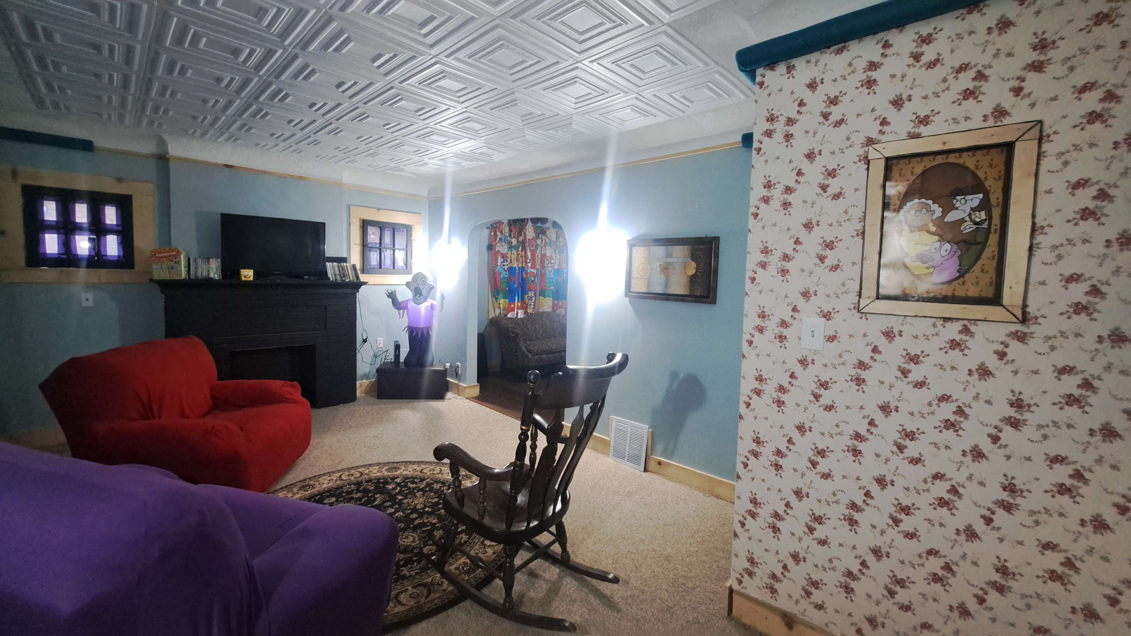 The image shows a living room with a red and purple sofa, a rocking chair, a fireplace, and floral wallpaper.