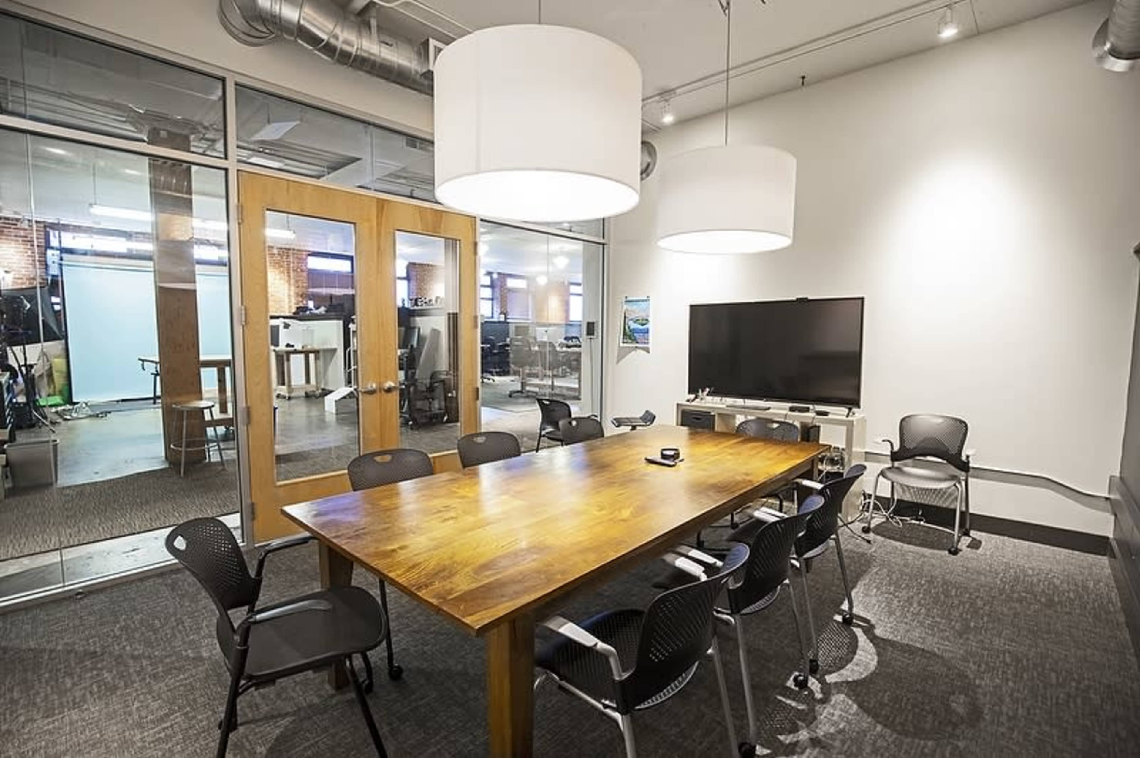 A modern meeting room features a large wooden table surrounded by black chairs, with two pendant lights hanging above and a wall-mounted TV at one end.