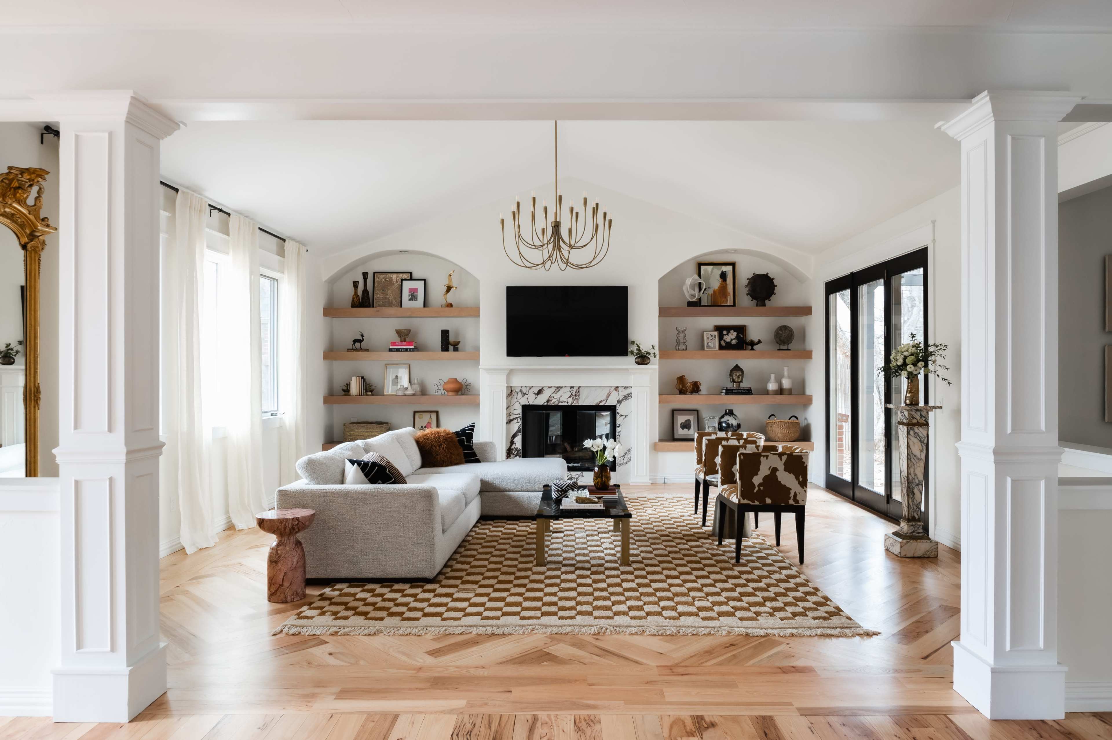 The image shows a spacious living room with a light-colored sofa, a checkered rug, a fireplace, and shelves displaying decorative items, all under a vaulted ceiling with a chandelier.