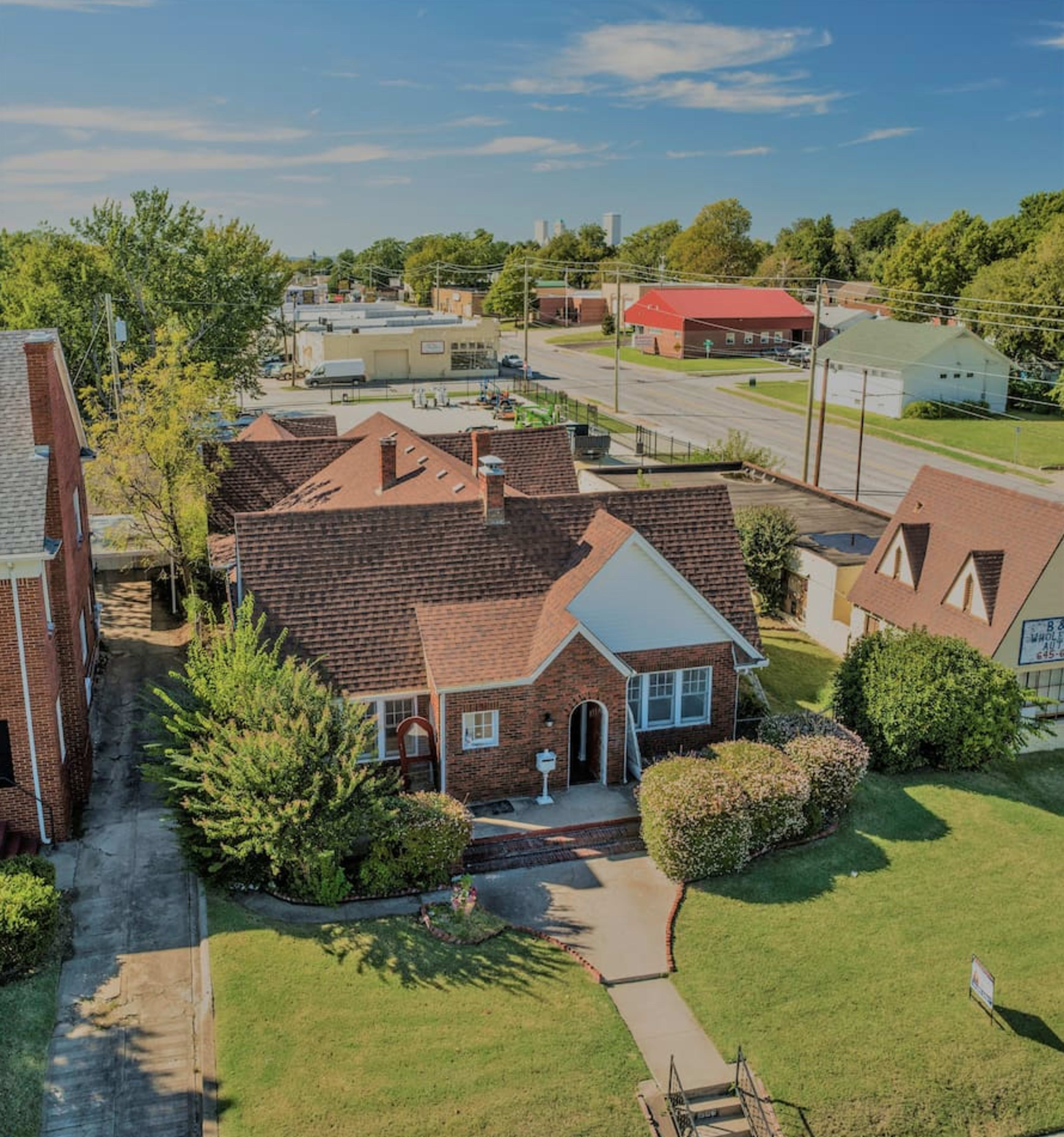 The image shows a red-brick house with a landscaped front yard and a paved pathway, situated near a street with several commercial buildings in the background.