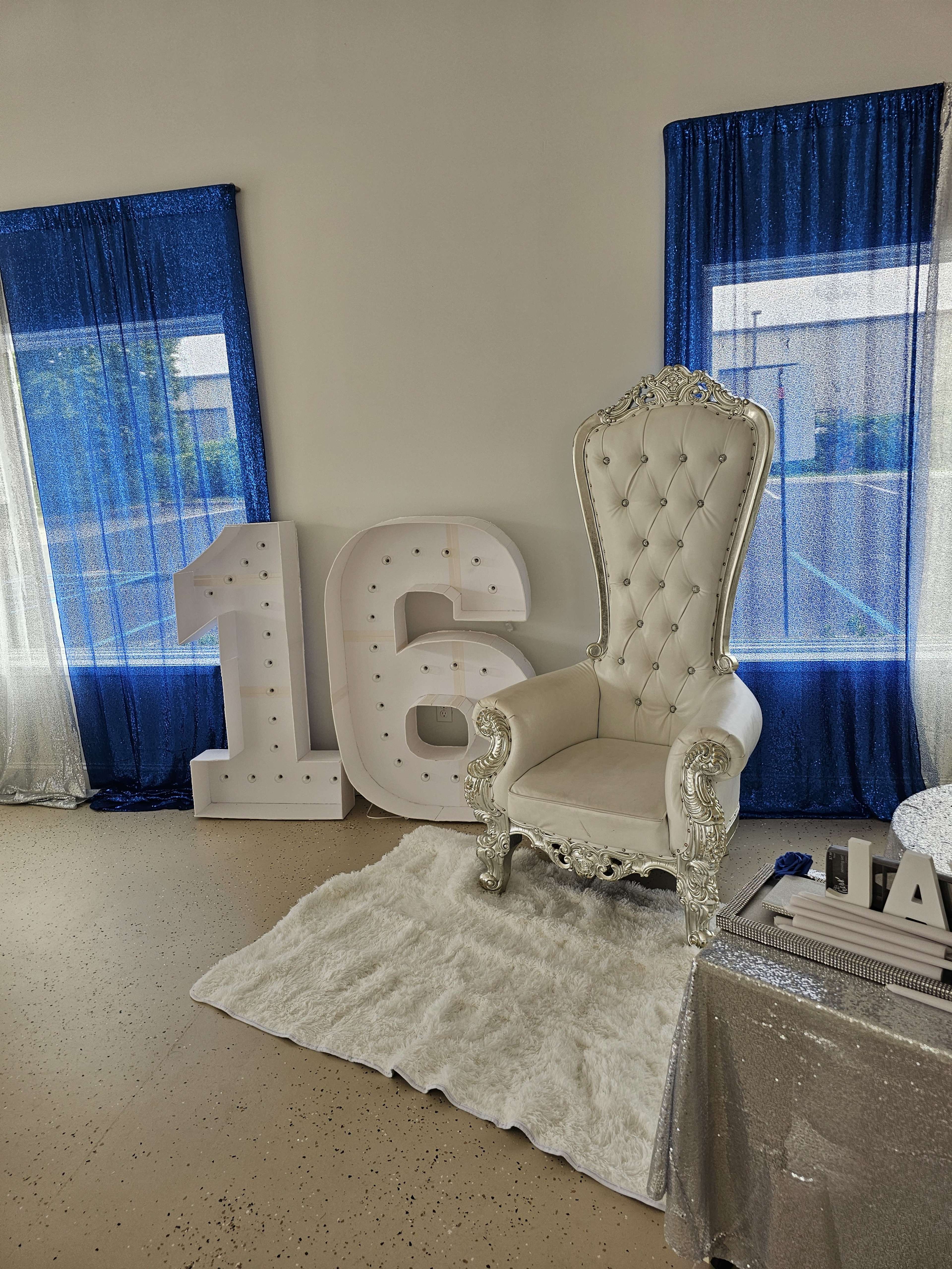A decorative setup for a sixteenth birthday celebration, featuring a white throne chair, large illuminated "16" numbers, and blue curtains.