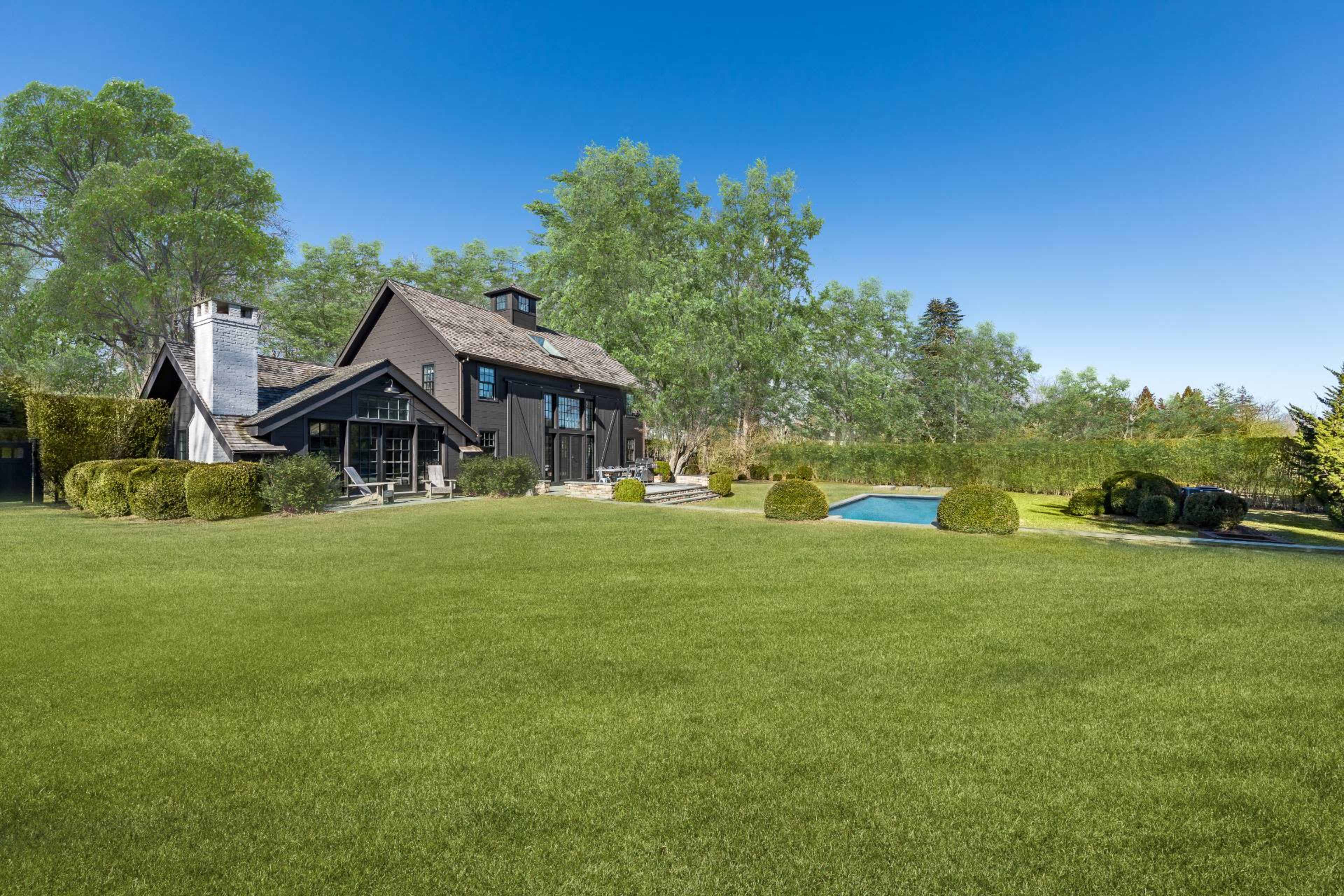 A black wooden house with a chimney, surrounded by a well-manicured lawn and a swimming pool in the backyard.