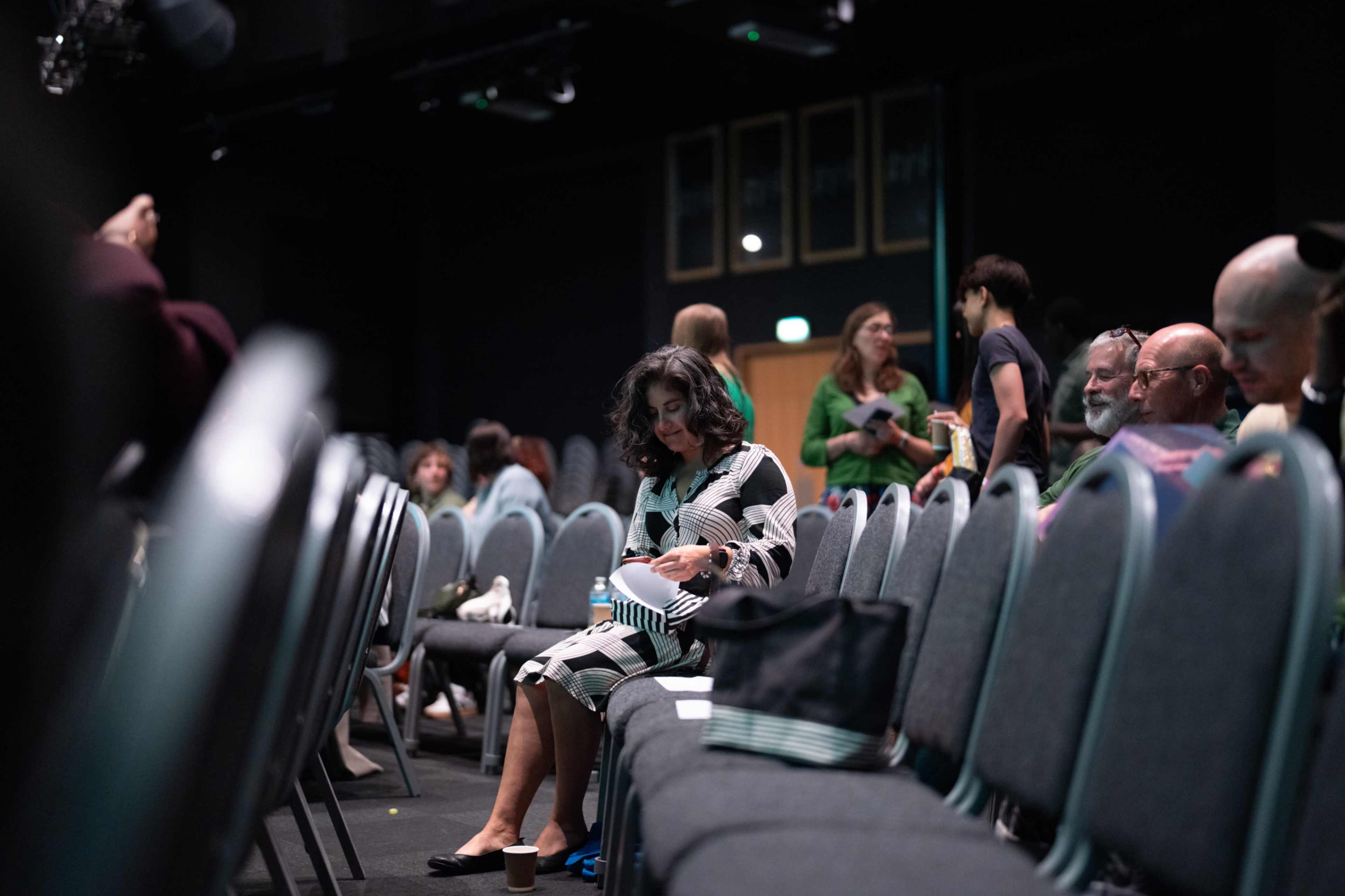 A woman sits alone in a row of empty chairs in a dimly lit auditorium while other attendees engage in conversation nearby.