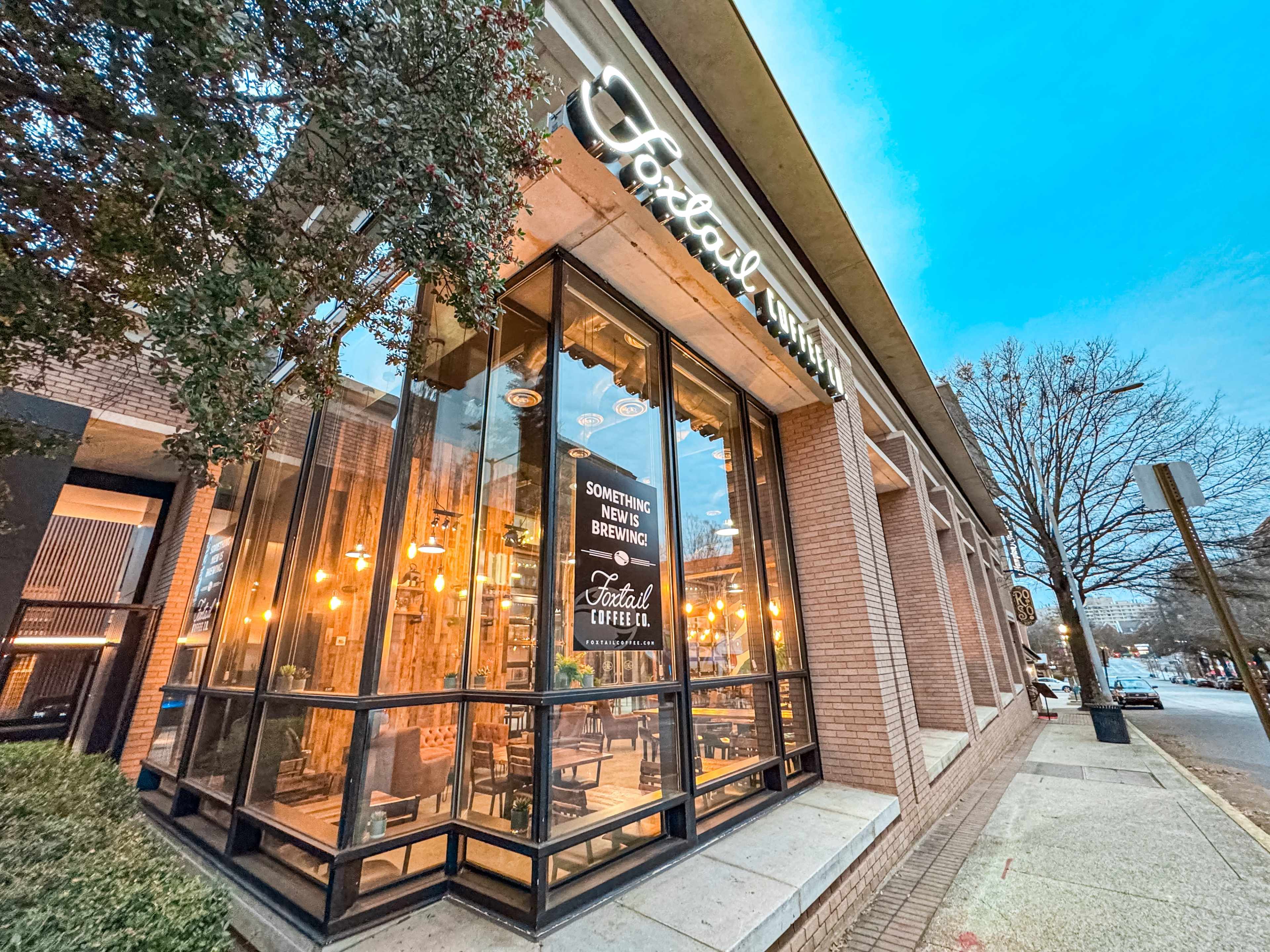 A coffee shop with large windows and a modern sign is located on a brick sidewalk, surrounded by trees and streetlights.
