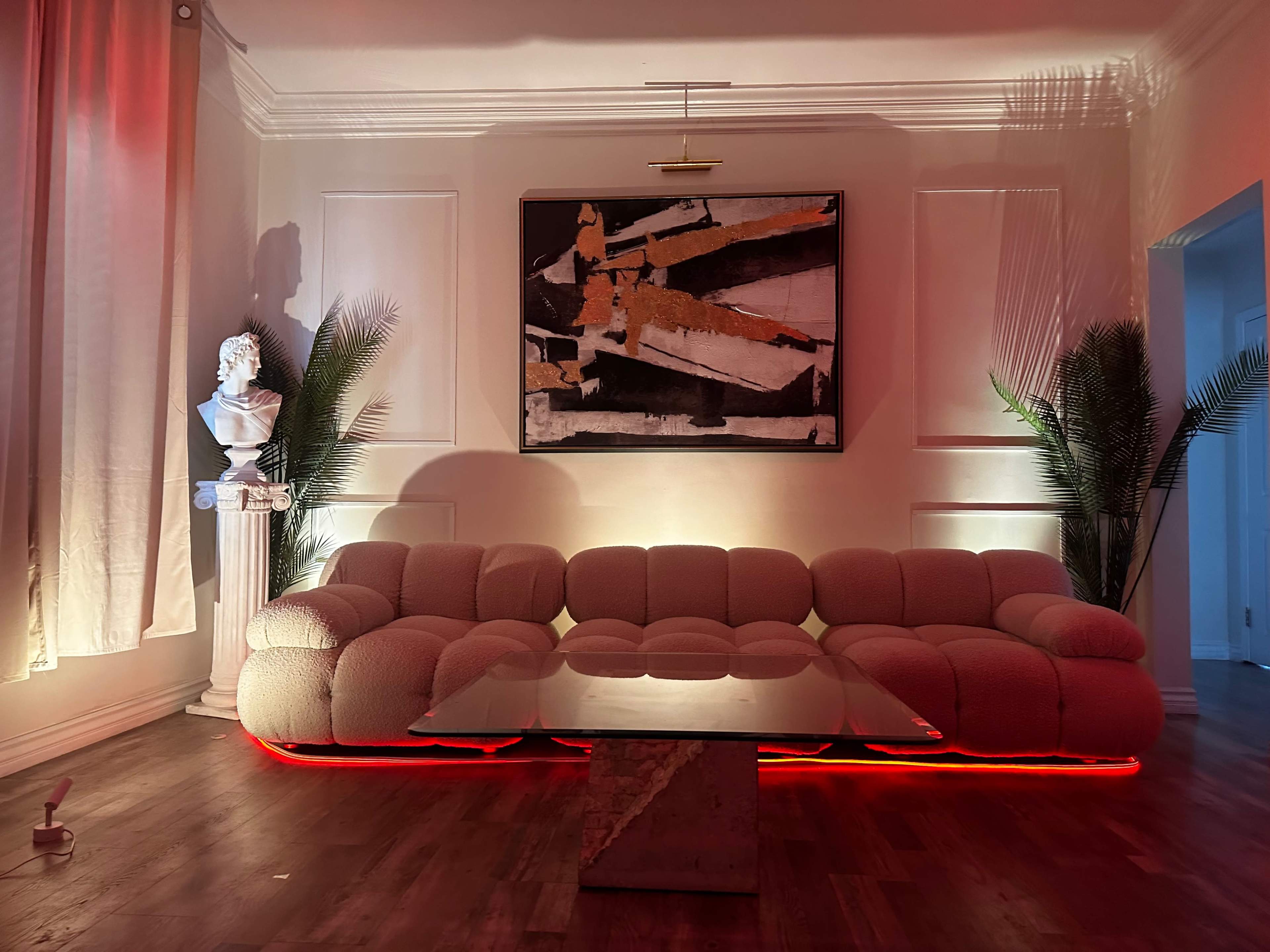 A modern living room features a curved, light-colored sofa, a glass coffee table, a large abstract painting on the wall, and indoor plants illuminated by soft lighting.
