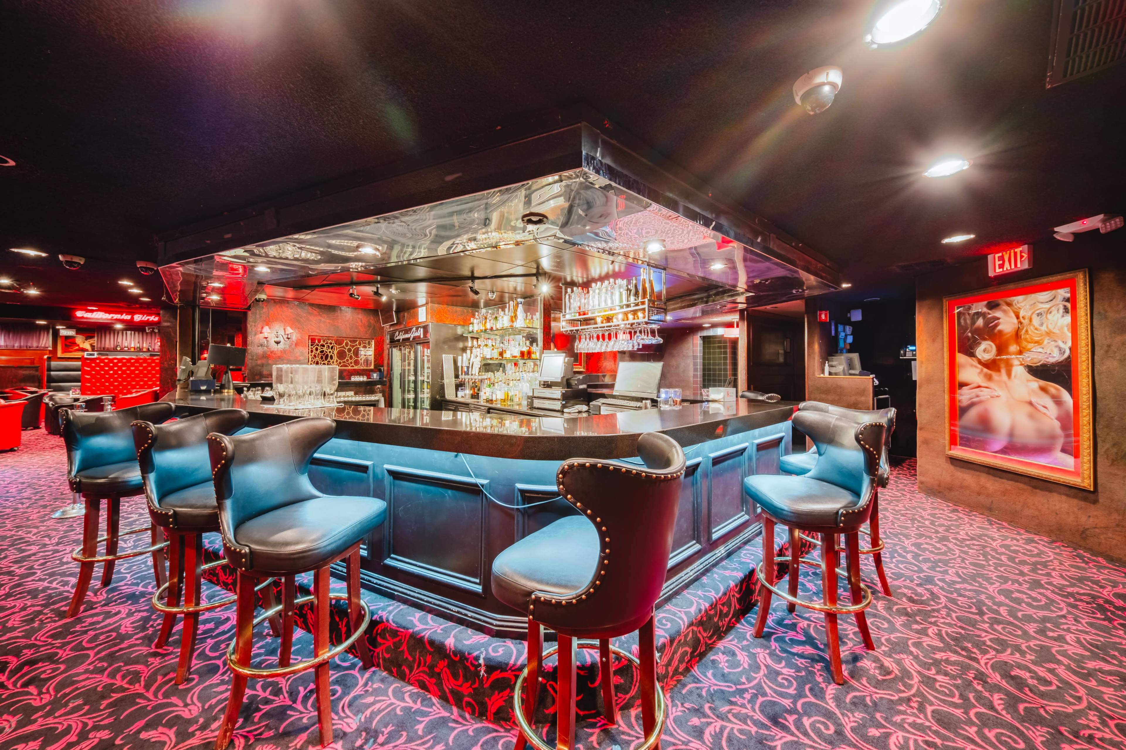 The image shows a dimly lit bar with a glossy counter, several high-backed stools, and shelves lined with bottles in a vibrant atmosphere.