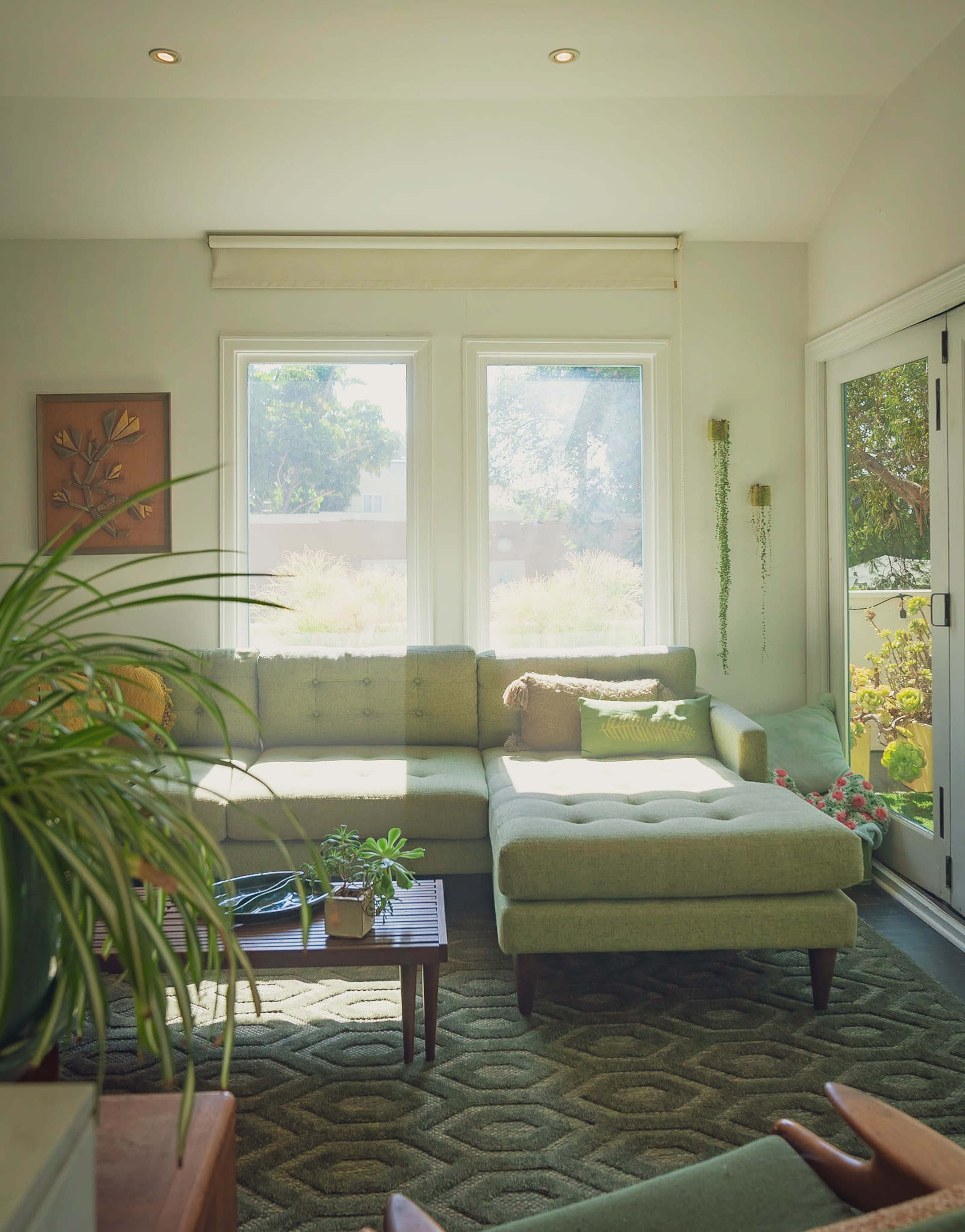 The image shows a bright living room with a green sectional sofa, a wooden coffee table, and large windows letting in natural light.