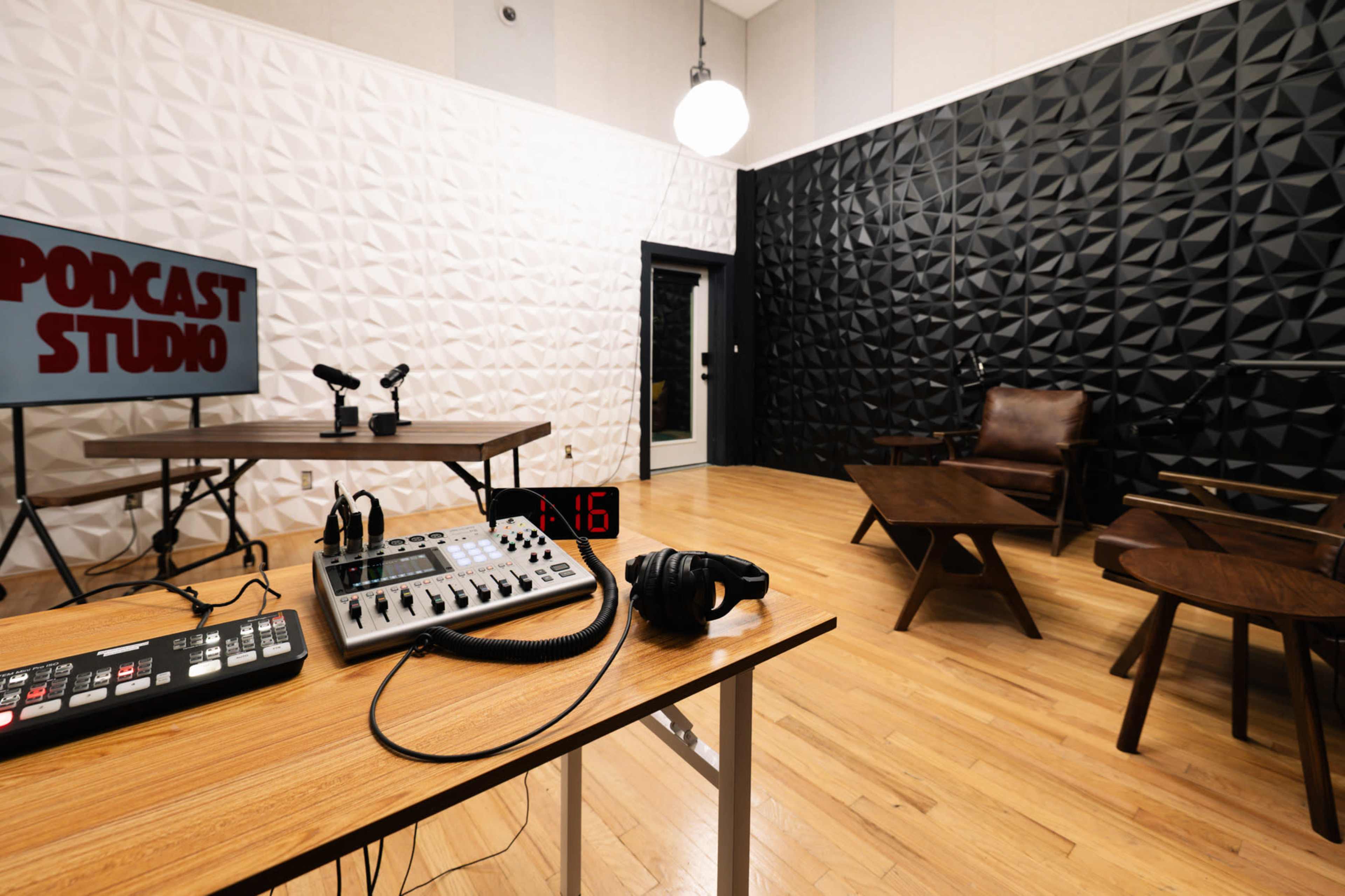 The image shows a podcast studio with a wooden table, a mixer, headphones, and two microphones positioned in front of a black and white geometric wall design.