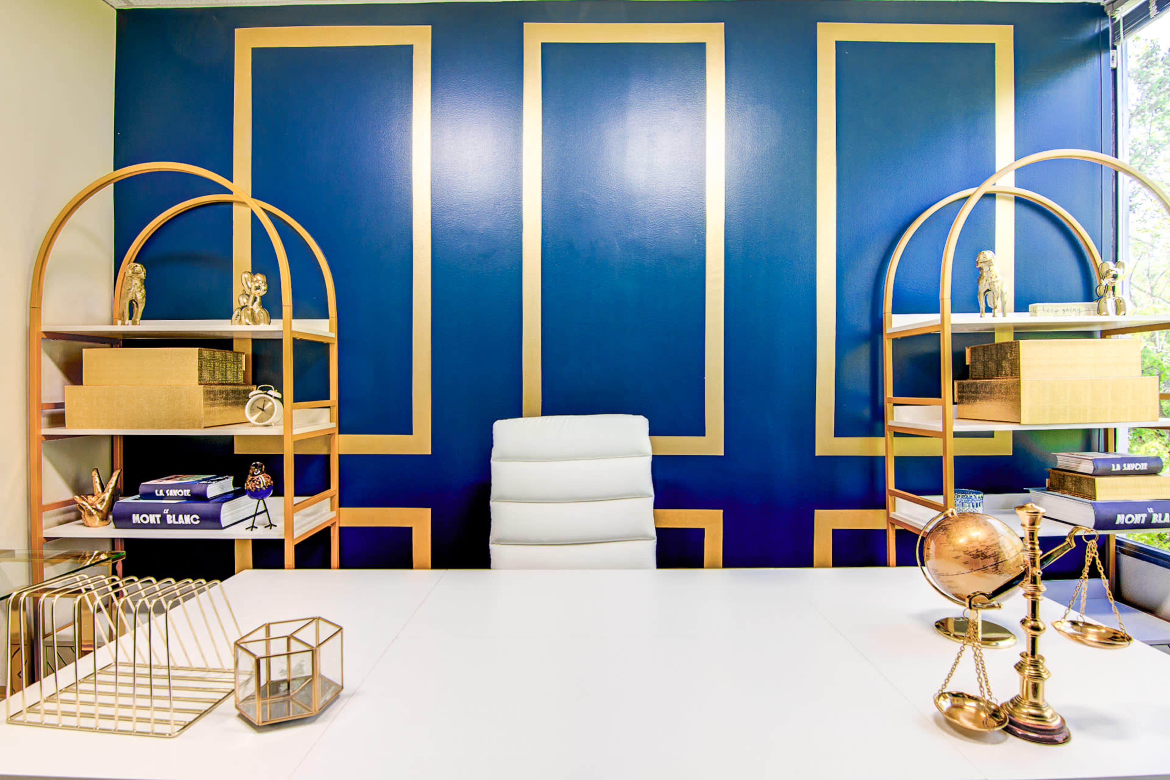 A modern office space features a white desk centered under a blue wall with gold geometric designs and shelves on either side displaying decorative items.