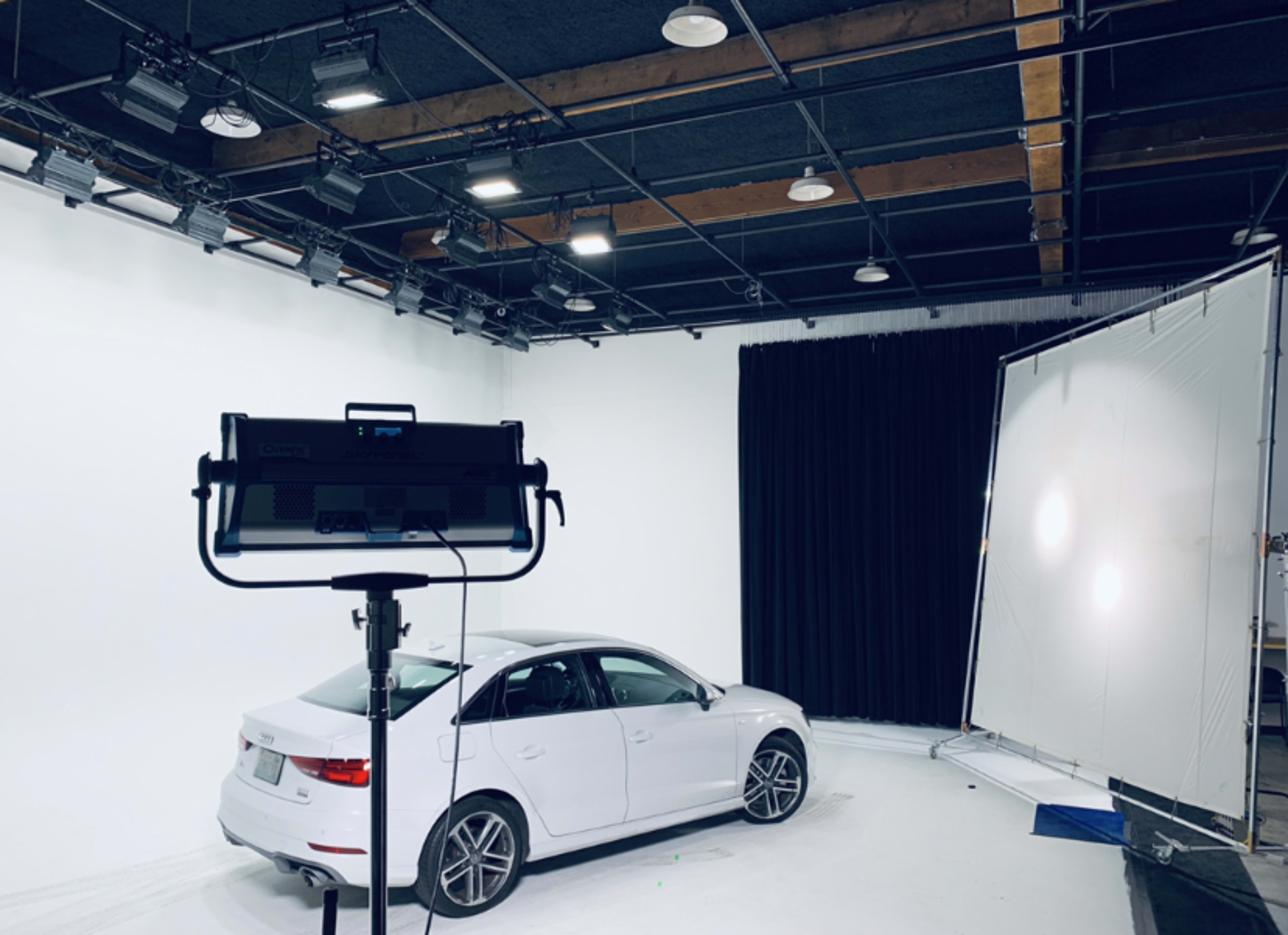 A white car is positioned in a well-lit studio with a black curtain in the background and lighting equipment visible in the foreground.