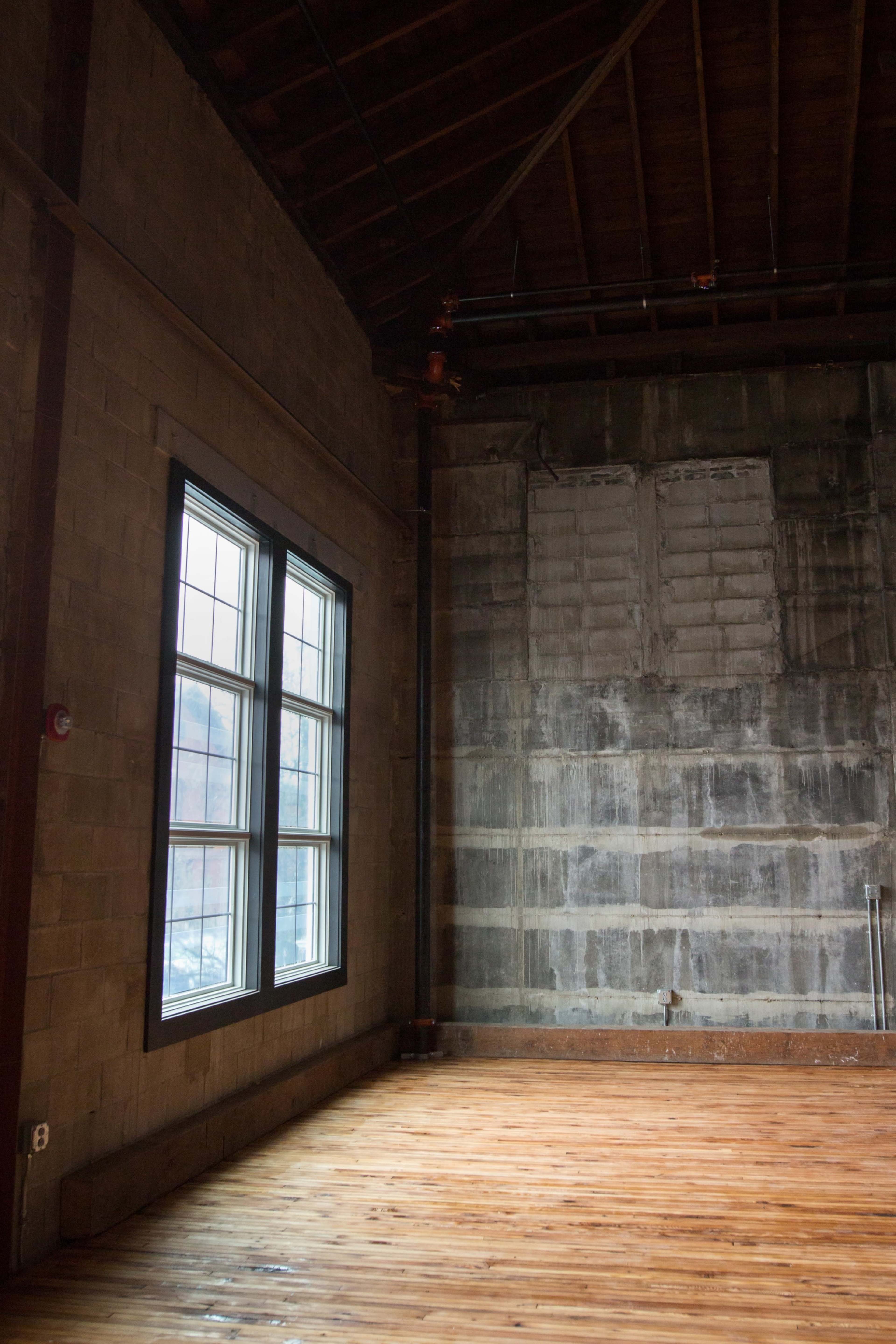The image shows an interior space with exposed wooden floors and a large window on a painted wall, adjacent to an unfinished concrete wall.