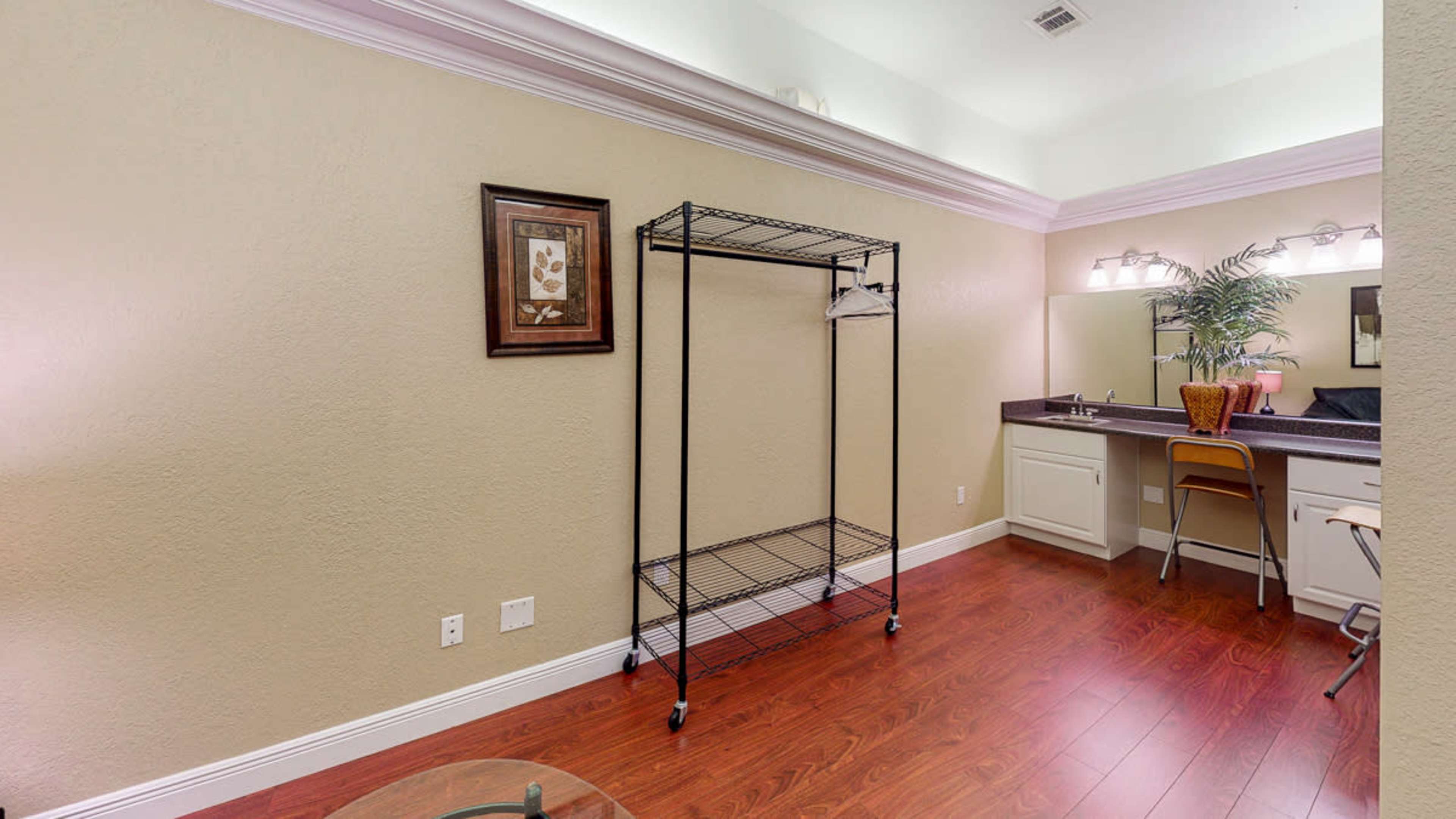The image shows an interior room with a wire clothing rack, a desk with a chair, and a mirror, all set against a light-colored wall and hardwood floor.