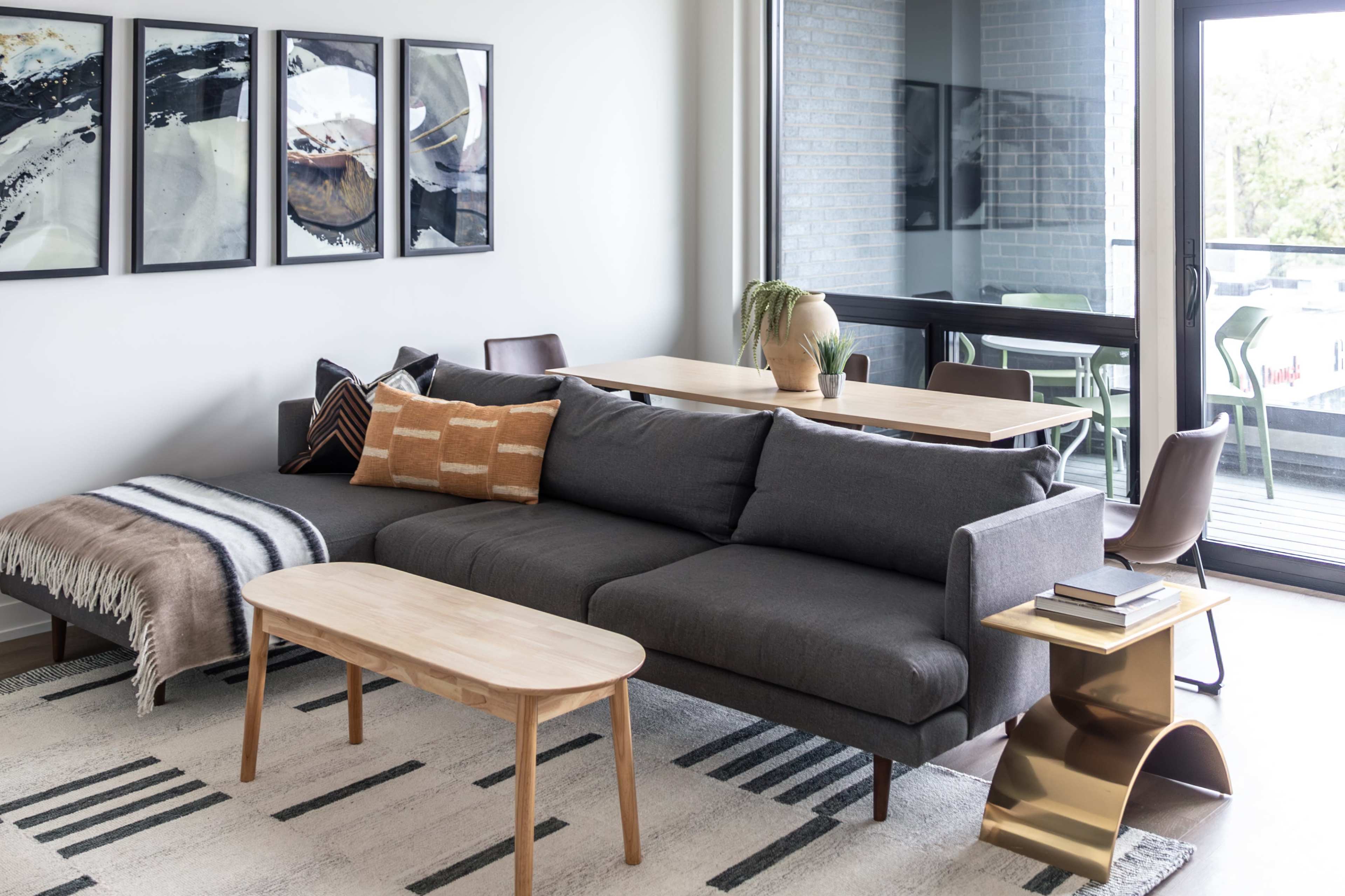 A modern living room features a gray sectional sofa, a wooden coffee table, and abstract art on the walls, with large windows providing outdoor views.