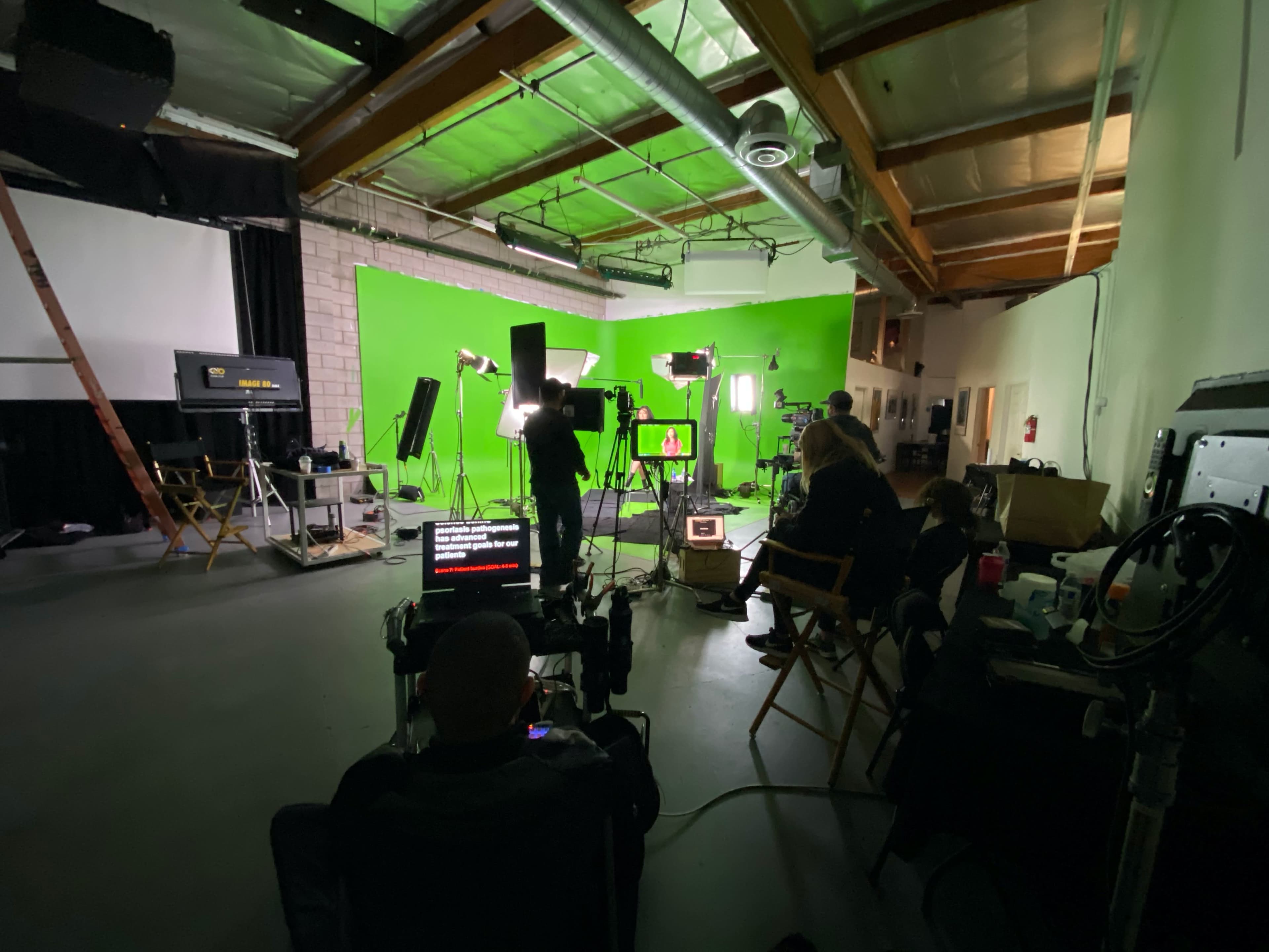 A film crew is set up in a studio with a green screen background, multiple lights, and equipment arranged for a shoot.
