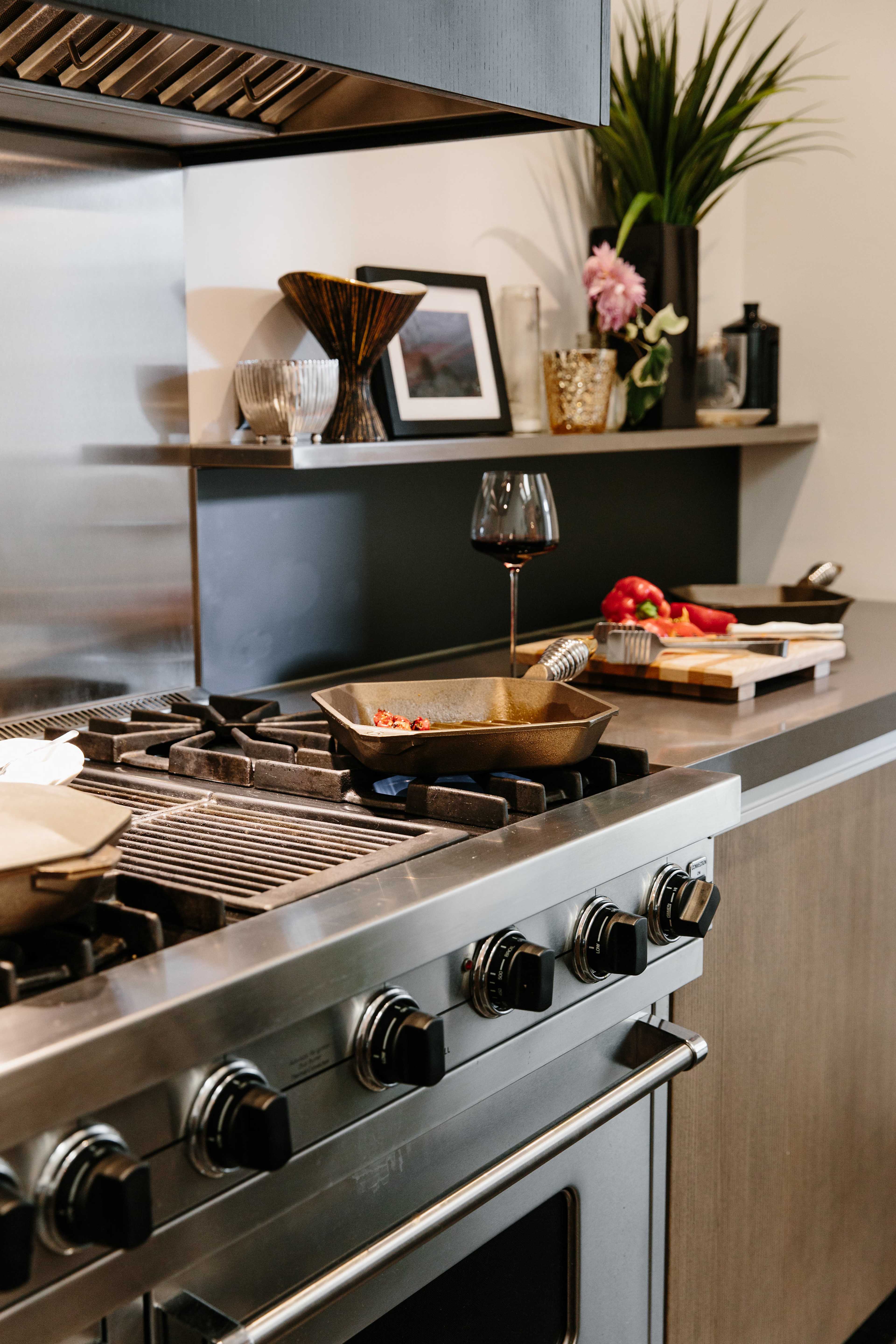 A modern kitchen features a gas stove with a cast iron pan and a glass of red wine on the countertop, alongside fresh vegetables and kitchen utensils.