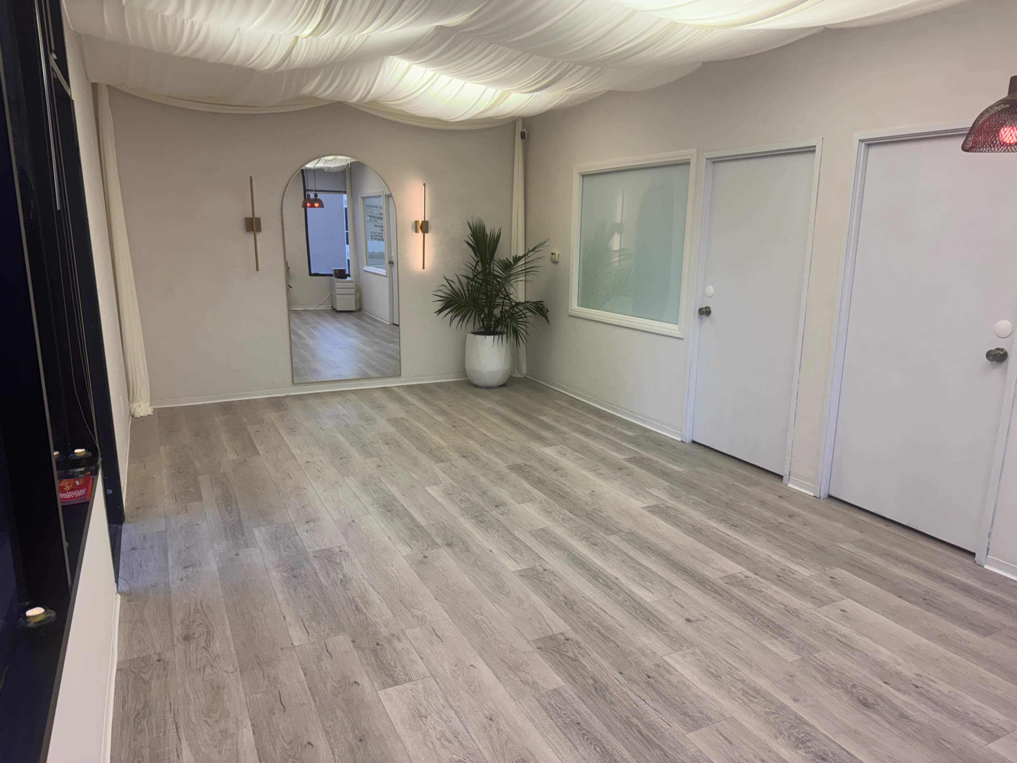 The image shows an empty, well-lit room with light wooden flooring, a large mirror on the wall, and two doors leading to other spaces.