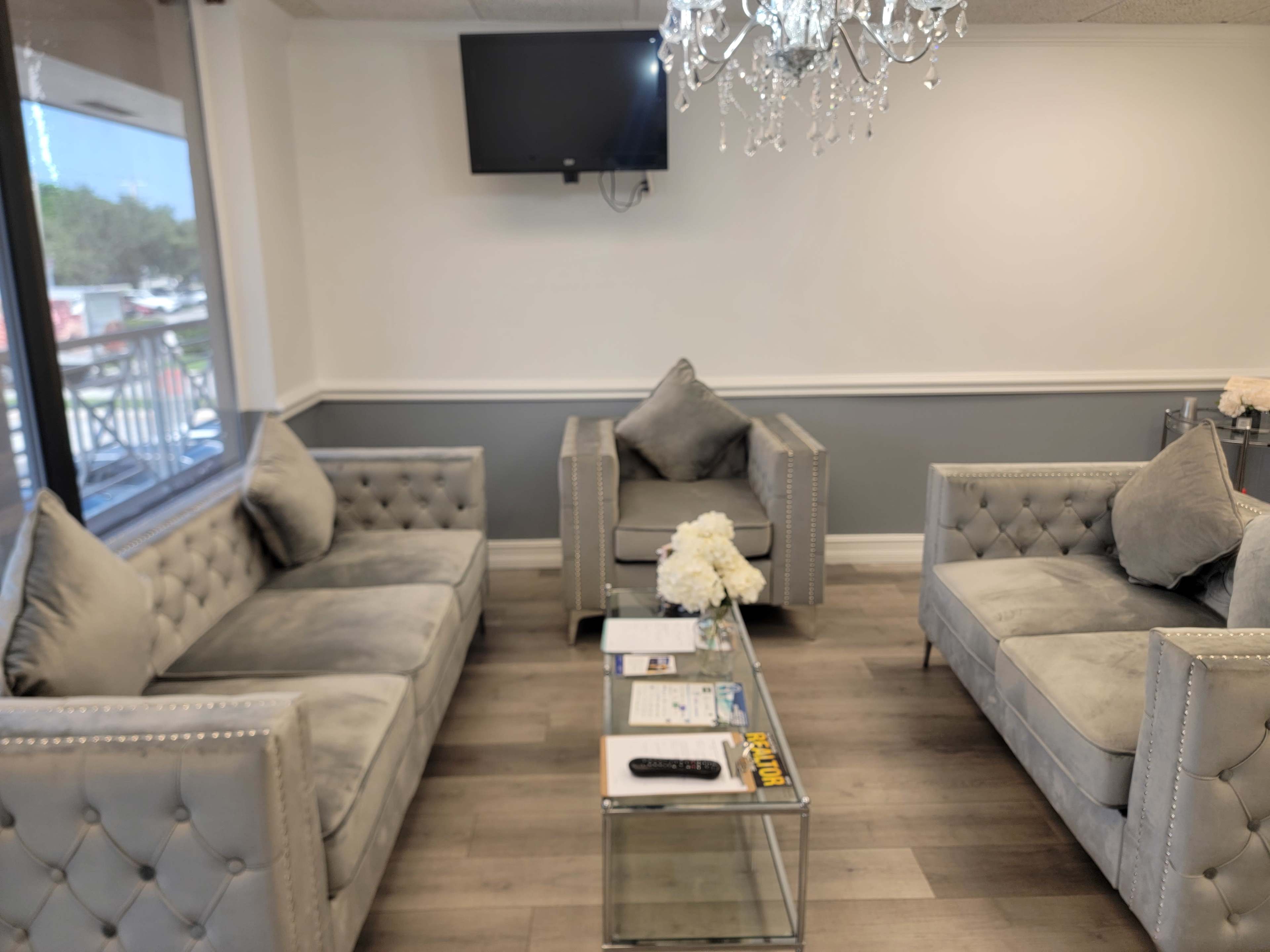 The image shows a modern waiting area with two tufted gray sofas, a glass coffee table, and a minimalist television mounted on the wall.