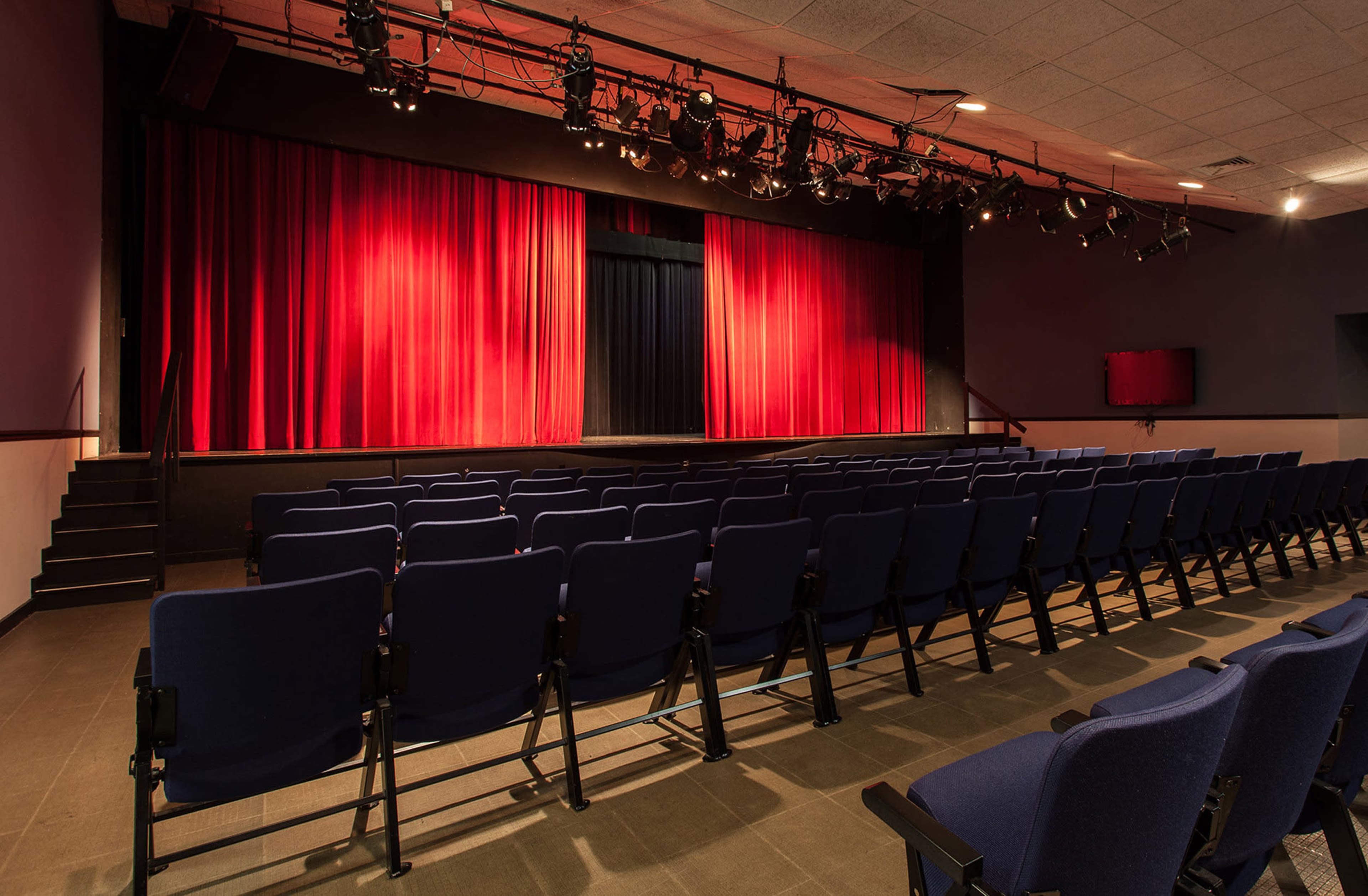 An empty auditorium with rows of blue seats facing a stage covered with red curtains.