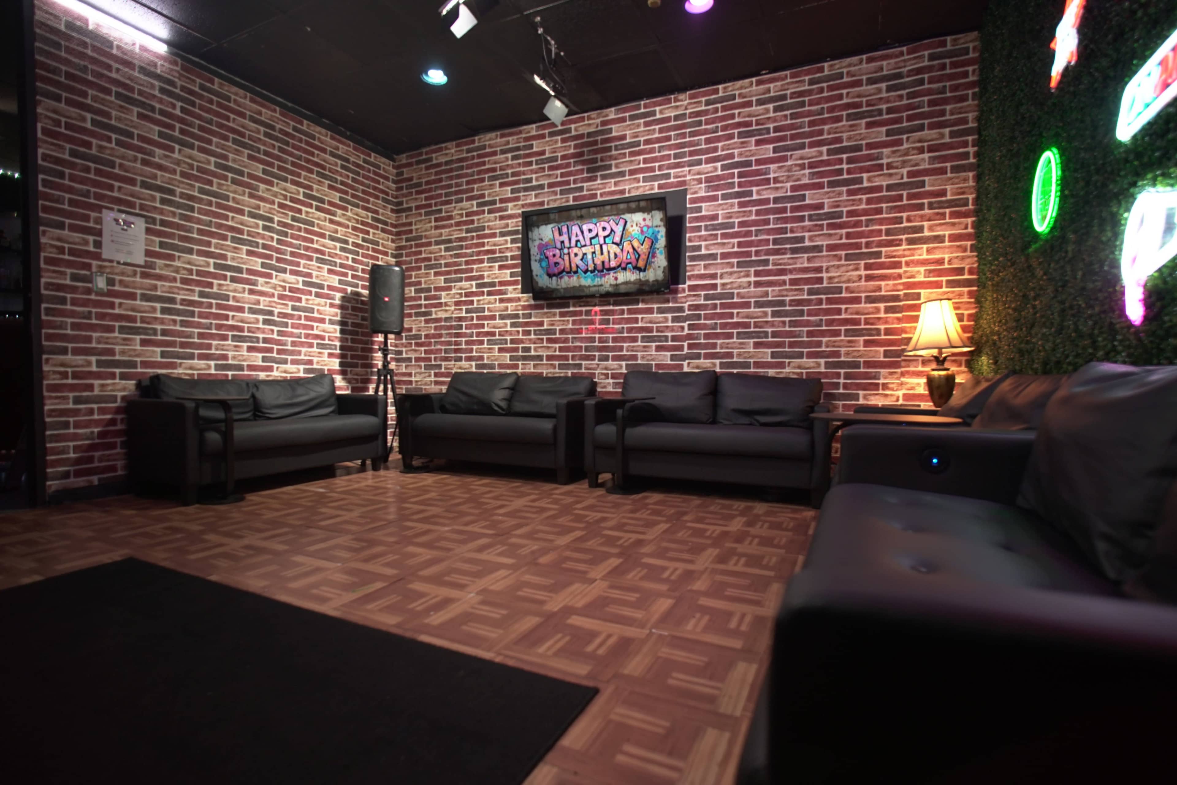 The scene shows a small gathering space with brick-patterned walls, several black couches arranged around a carpet, and a "Happy Birthday" sign displayed on a screen.