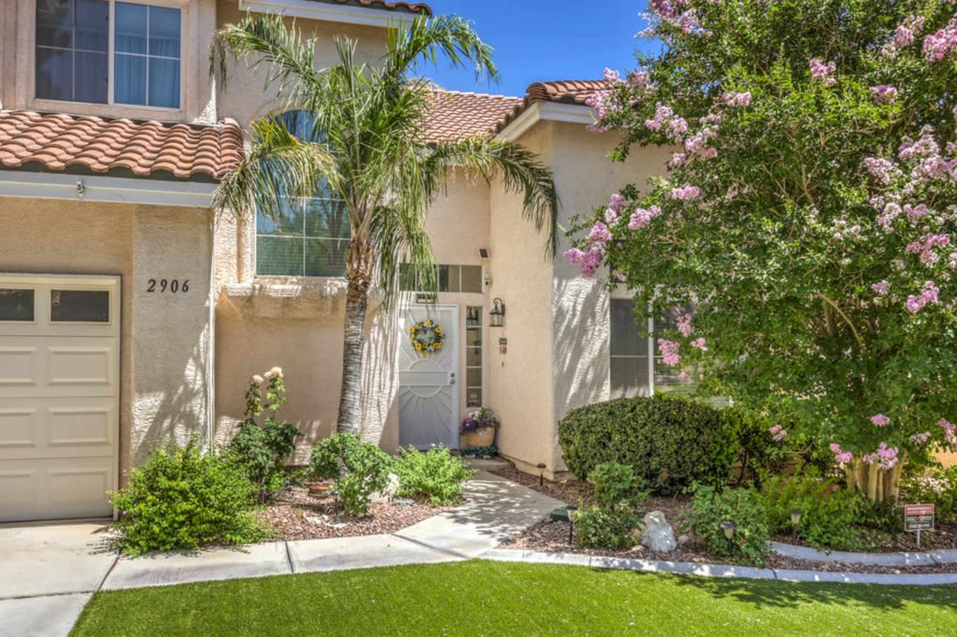 The image shows a home with a beige exterior, a tile roof, and a well-kept garden featuring palm trees and flowering shrubs.
